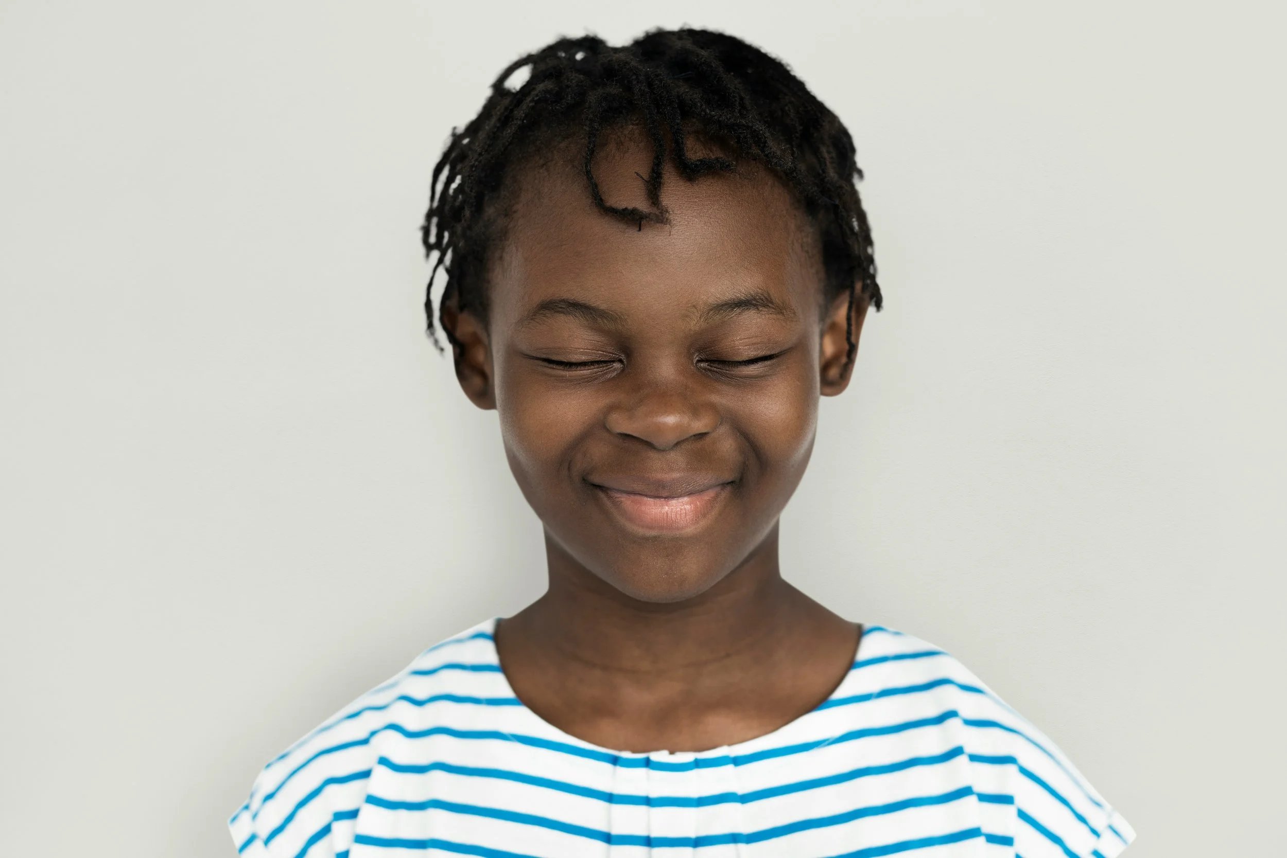 Smiling young boy with natural dreadlocks wearing a striped shirt against a plain white wall.