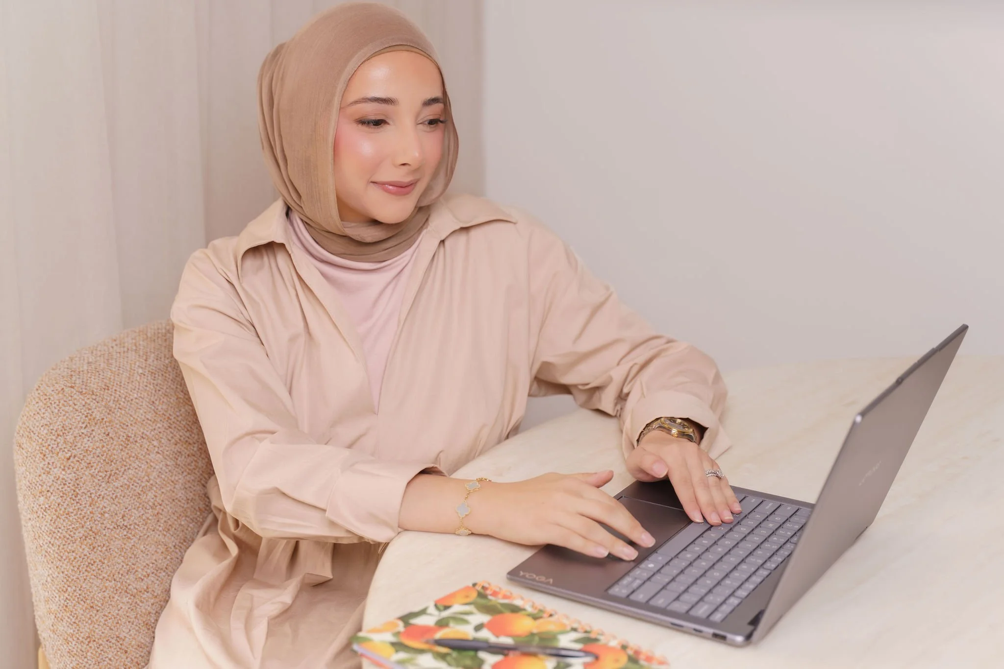 A young woman wearing a beige hijab and beige blouse sitting at a table, using a laptop with a colorful notebook and floral planner nearby.