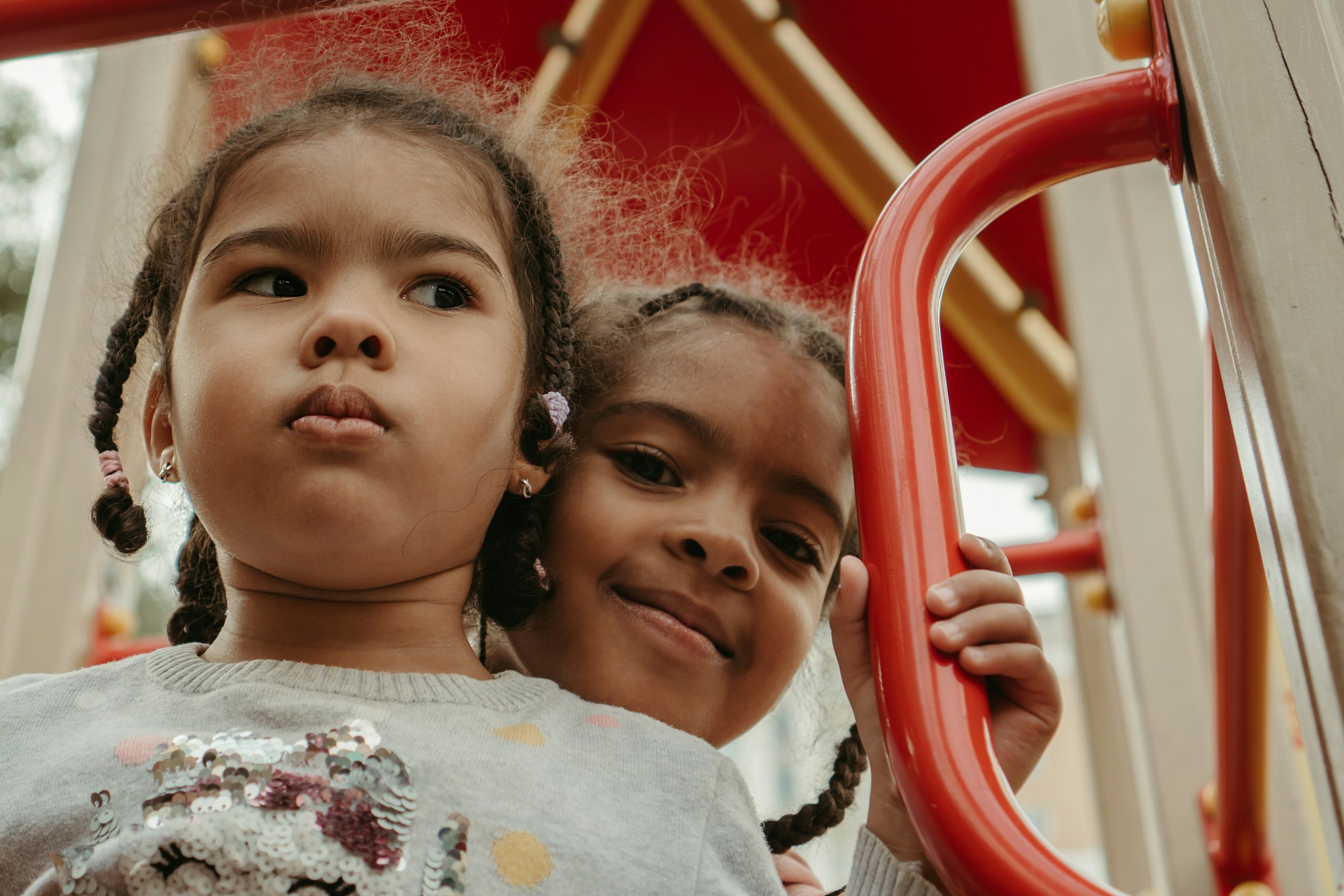 Two young girls with braided hair smiling, one holding onto a red playground structure.