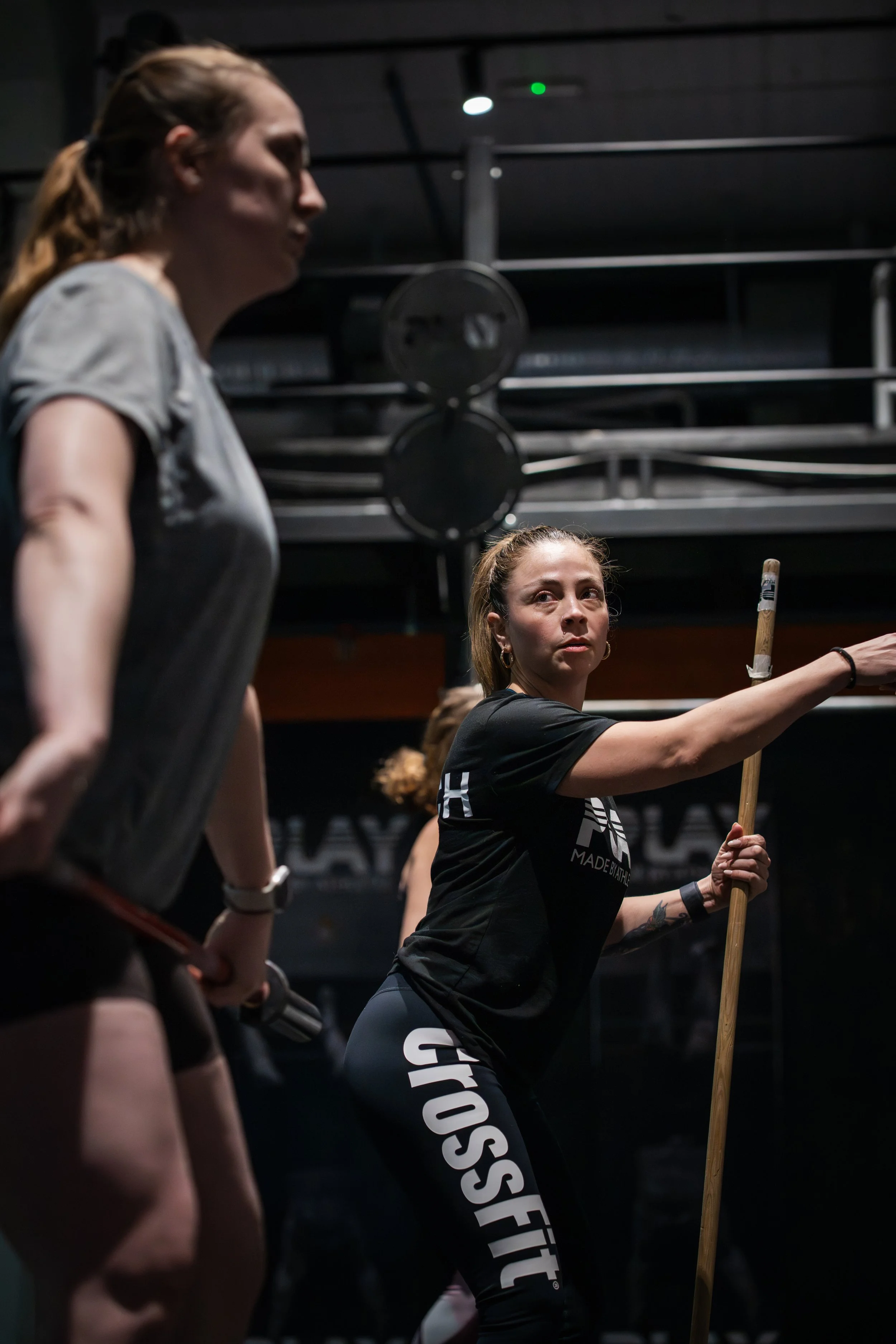 Two women are in a CrossFit gym. The woman on the right is holding a wooden pole, appearing to be instructing or demonstrating, while the woman on the left is approximately listening. The background shows gym equipment and a dark environment.