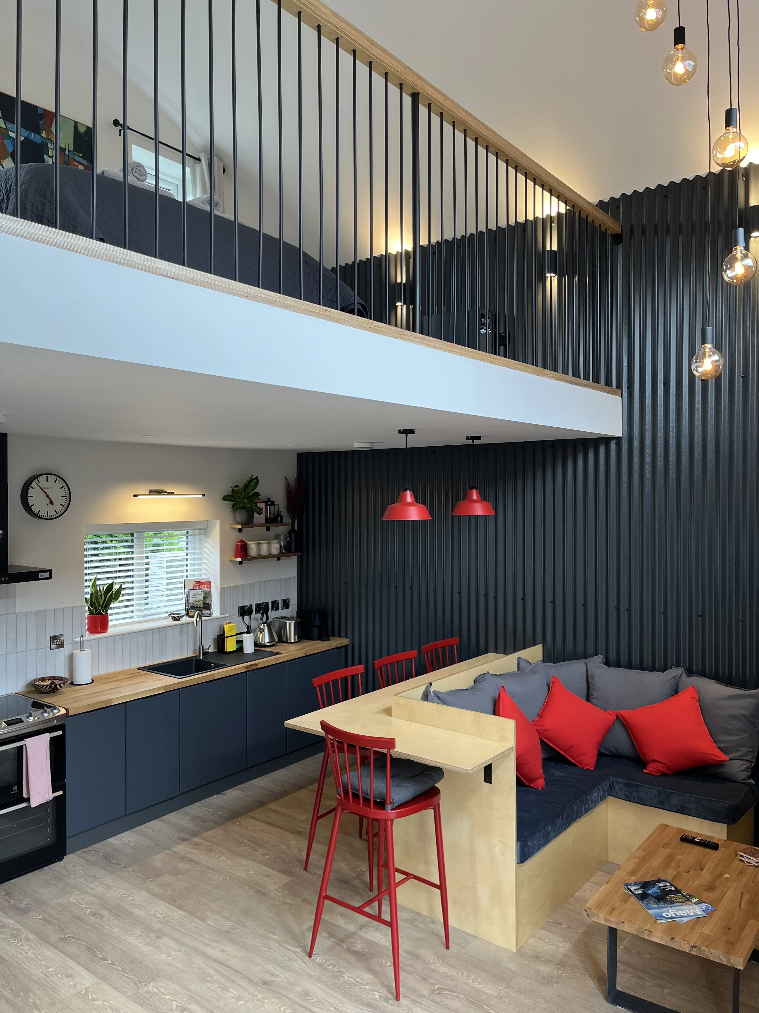Modern open-plan living space with a kitchen, dining area, and a mezzanine from above. The kitchen has dark blue cabinets, a wooden countertop, and white wall paneling. The dining area has a wooden table with red chairs and a built-in corner sofa wit