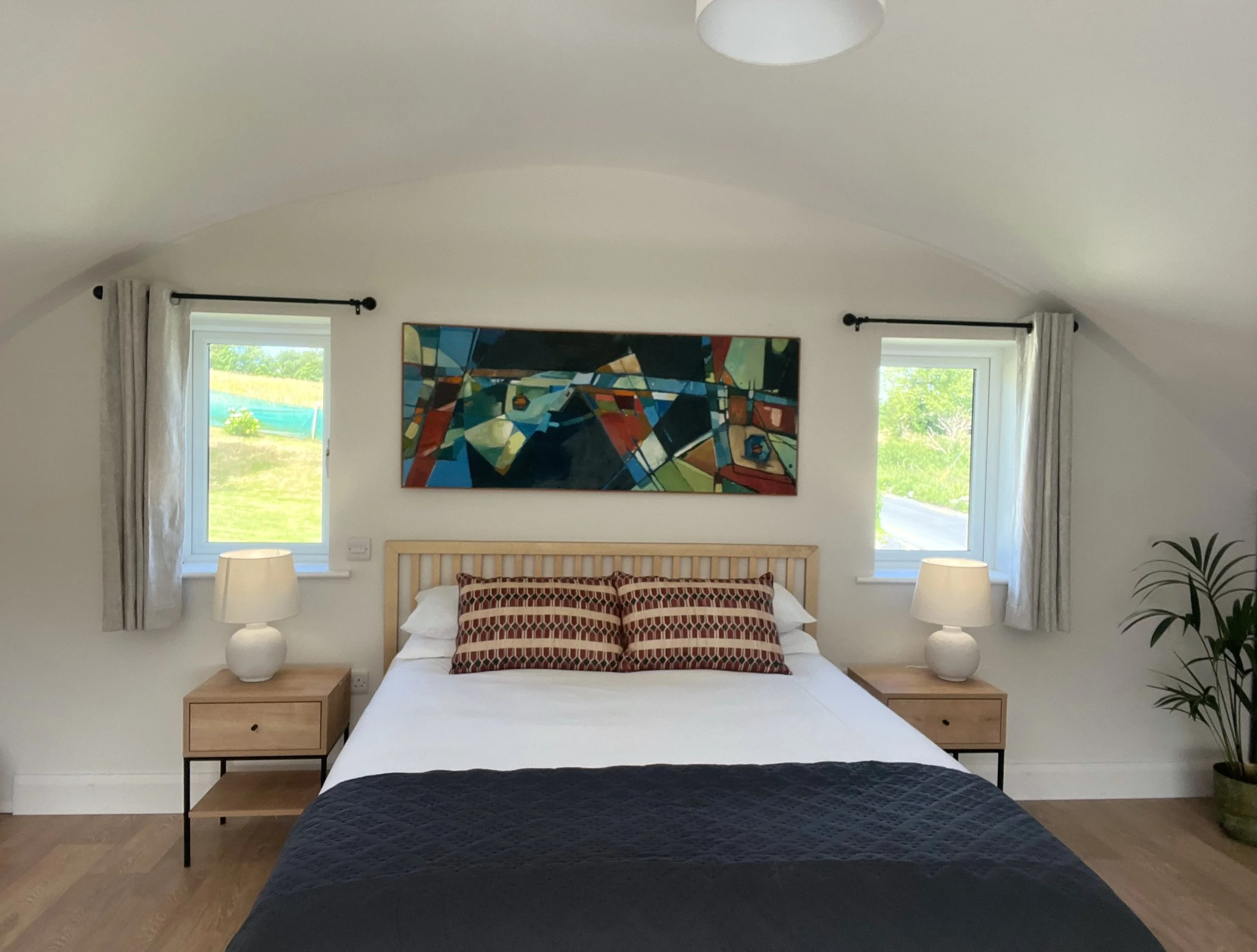 A bedroom with a large bed with patterned pillows, wooden nightstands with white lamps, two windows with curtains, a colorful abstract painting above the bed, and a potted plant to the right.