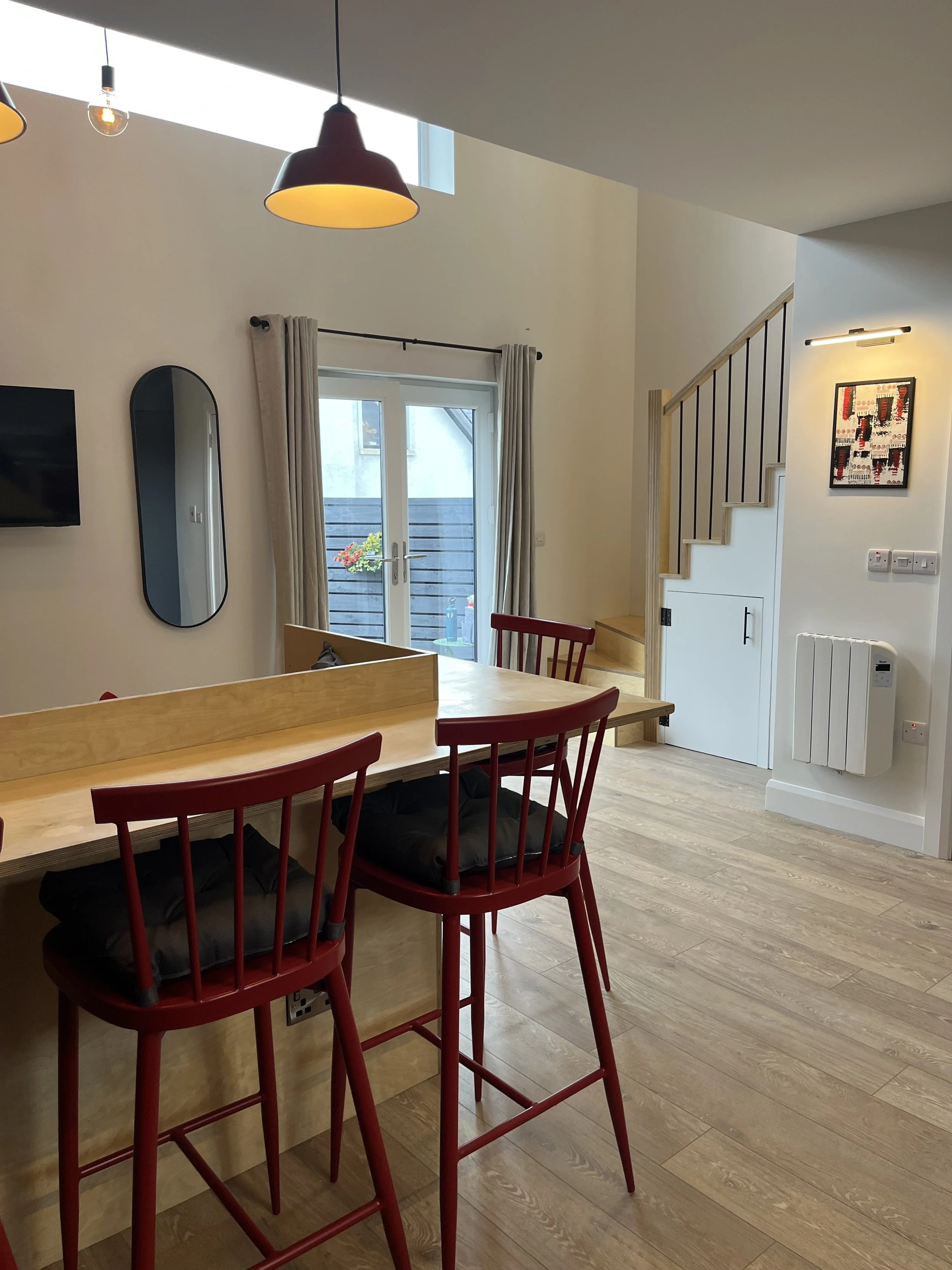 A dining area with a wooden table and red chairs, a staircase with black balusters, white walls, a glass door leading to a small balcony with plants, beige curtains, a wall-mounted TV, and various lights.