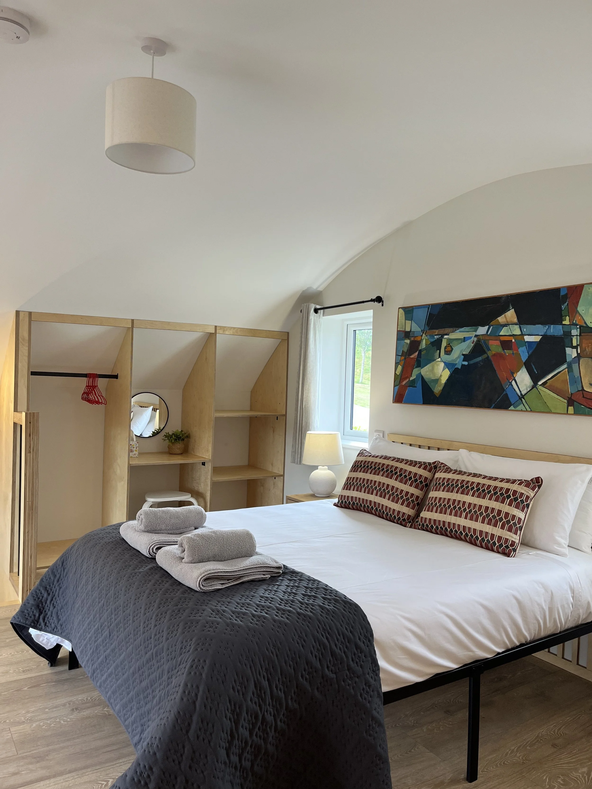 A cozy bedroom with a neatly made bed featuring white sheets and decorative pillows. Towels are folded at the foot of the bed. There's a wooden shelving unit with a small plant, a mirror, and a red hanger. A window with a curtain, a bedside table wit