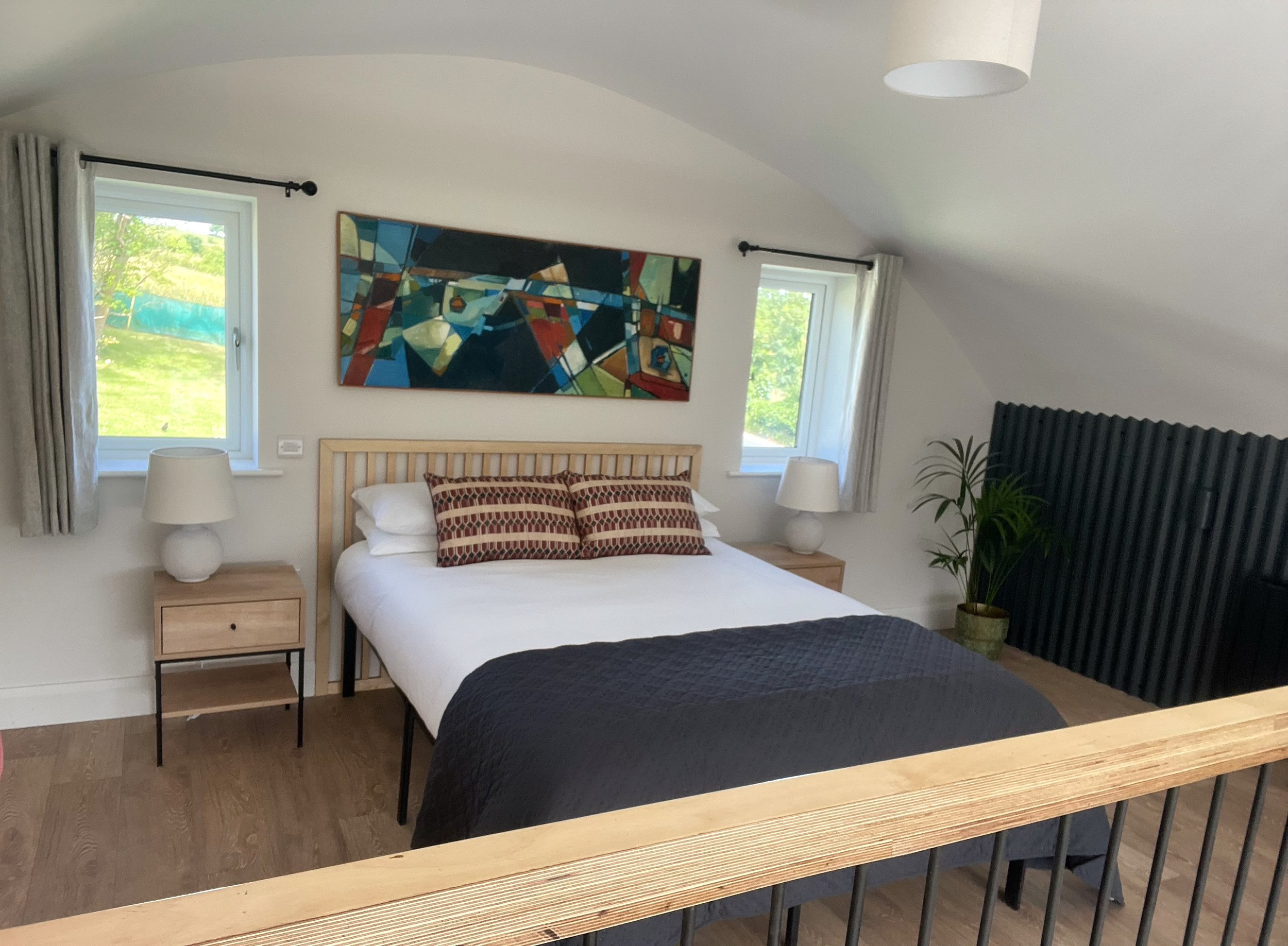 A bedroom with a bed, two windows with curtains, and modern artwork on the wall. There are two bedside tables with lamps, a plant in a pot, and a black radiator. A wooden railing is visible in the foreground.