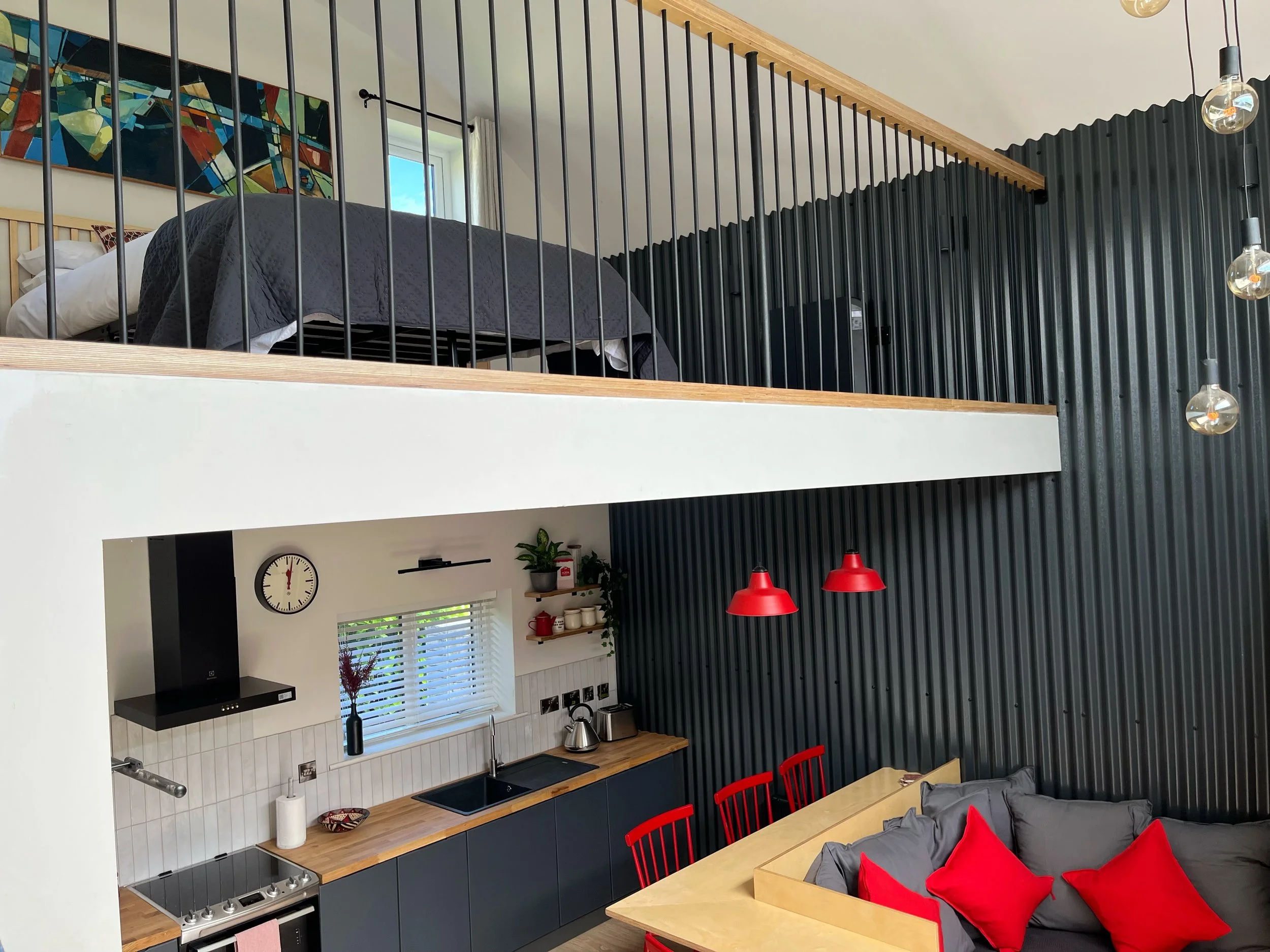 Open-plan kitchen and dining area with black and wood cabinetry, a white tiled backsplash, a black range hood, and red dining chairs. A window with white blinds, wall clock, and hanging light bulbs are visible. A loft bedroom with a bed, gray quilt, and a colorful abstract painting are seen above the kitchen.