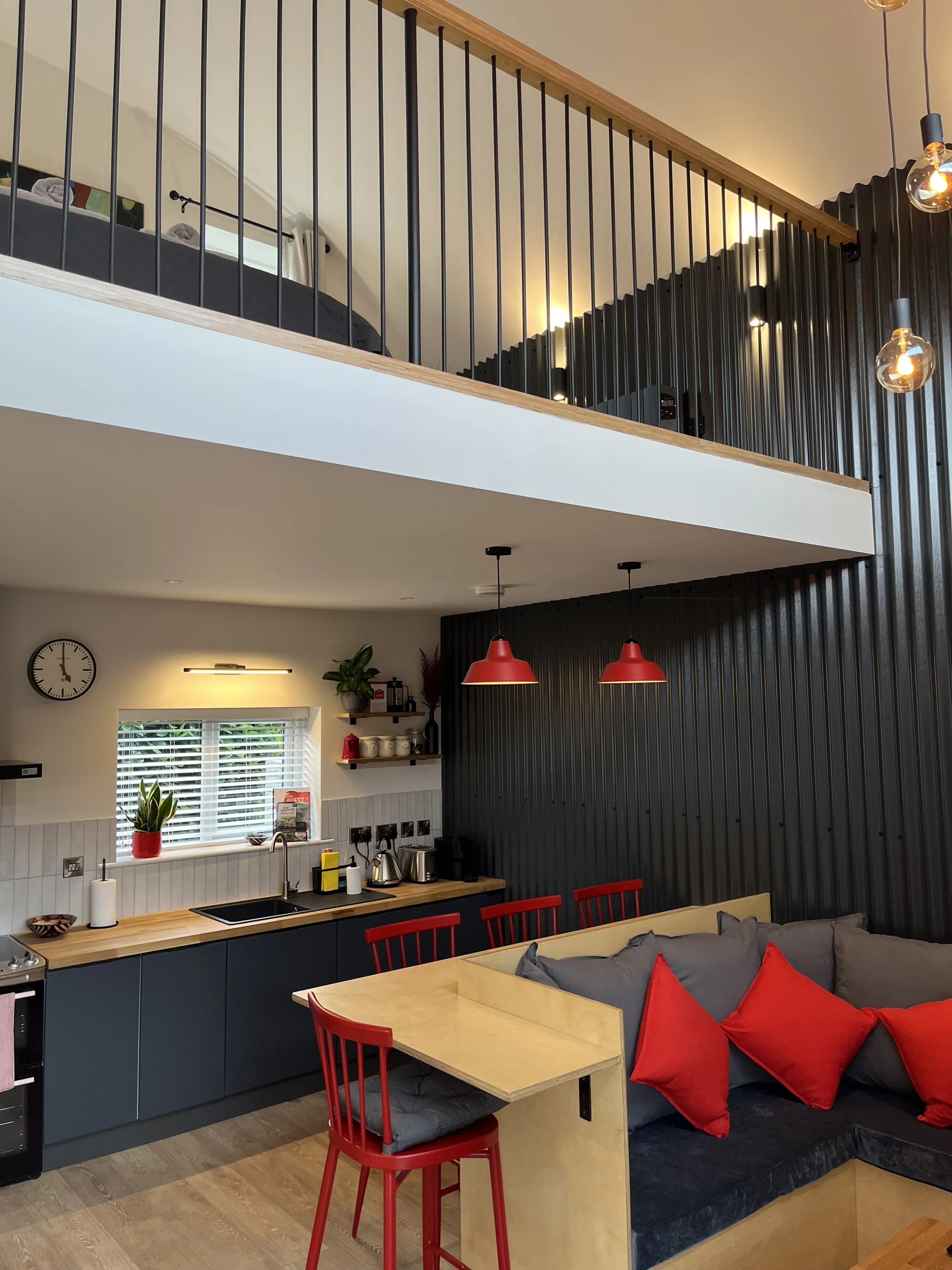 Modern kitchen and living area with black and red color accents, pendant lights, and a mezzanine with a bed.