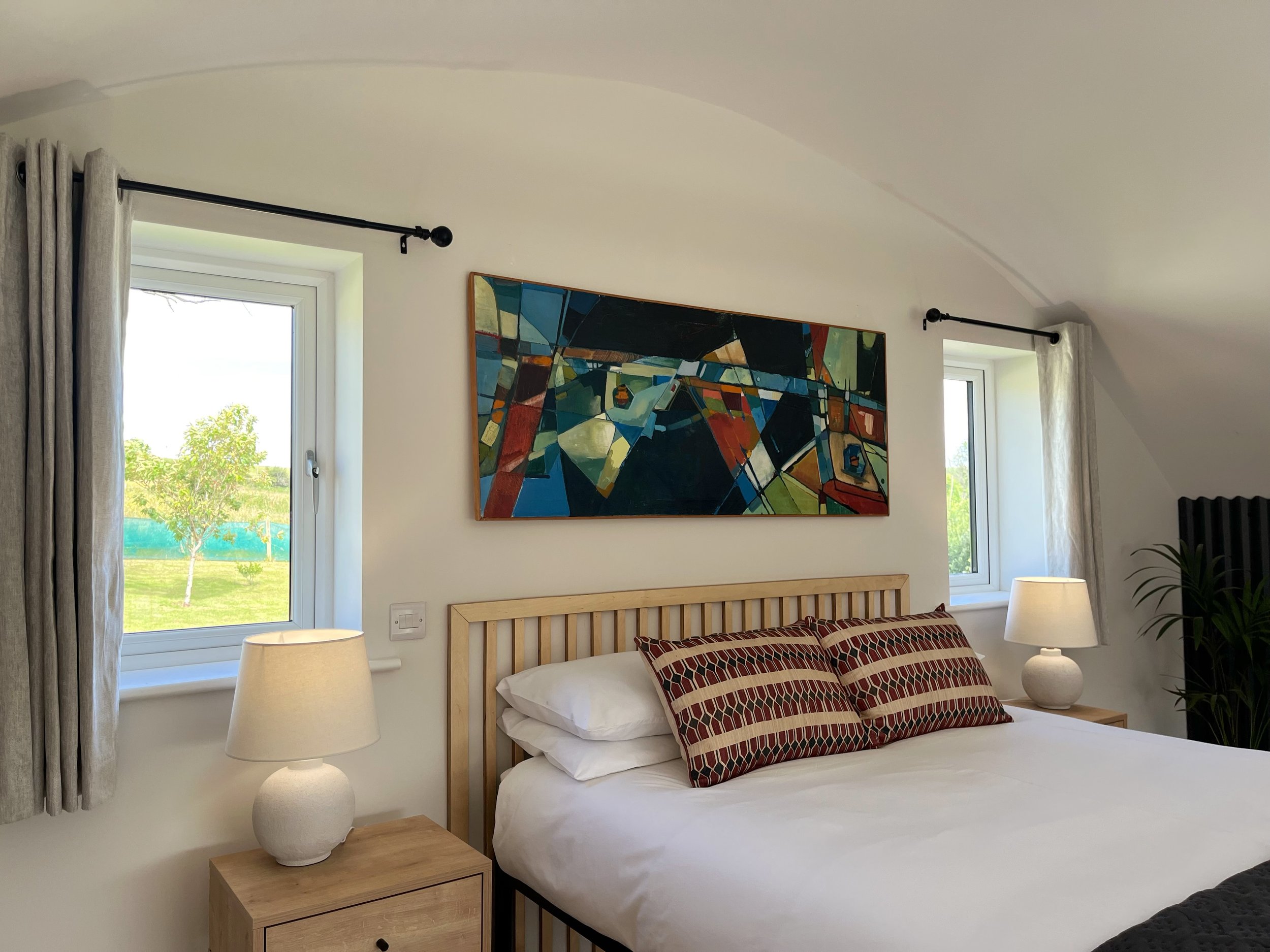 A cozy bedroom with a wooden bed frame, colorful striped pillows, and two large windows showing a green landscape outside. There are beige curtains, a nightstand with a white ceramic lamp, a modern abstract painting on the wall, and a small potted pl