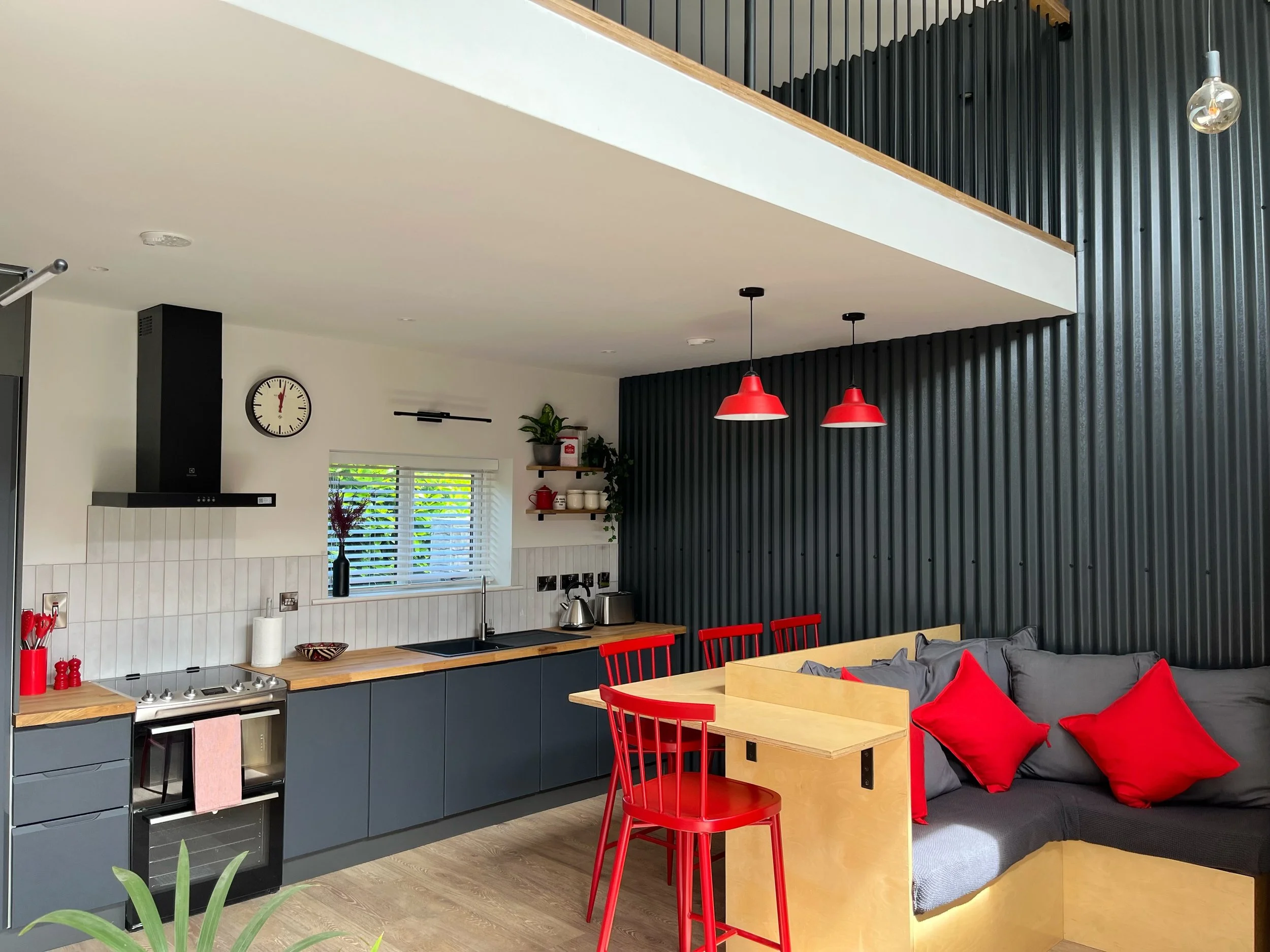 A modern kitchen with black corrugated metal wall, white ceiling, red pendant lights, and a dining area with red chairs and gray cushions with red pillows.