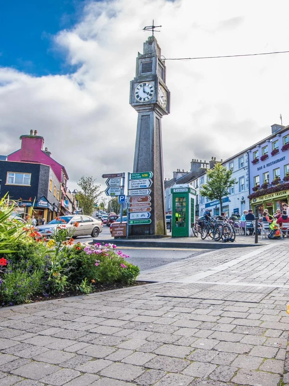 A town square with a tall clock tower at the center. There are colorful buildings, bicycles, a green phone booth, and a group of people sitting outside a bar called "The Clock Tavern." Flowers and plants are in the foreground, and signs with directions are attached to the clock tower.