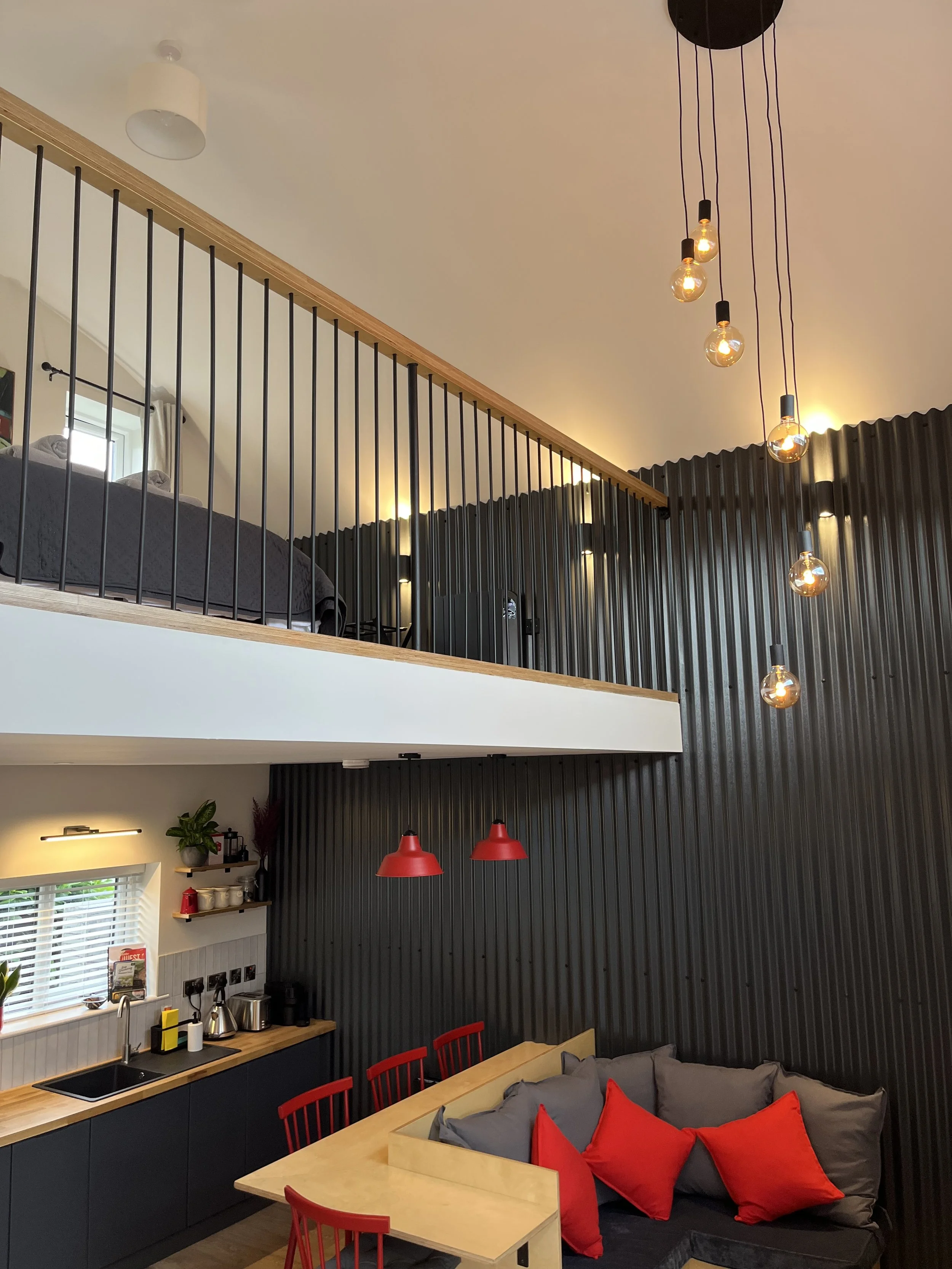 Interior view of a modern open-plan kitchen and living area with black paneled walls, a loft with black metal railing, wood accents, and pendant lighting. The kitchen has a small window with white blinds, black lower cabinets, a wooden countertop, an