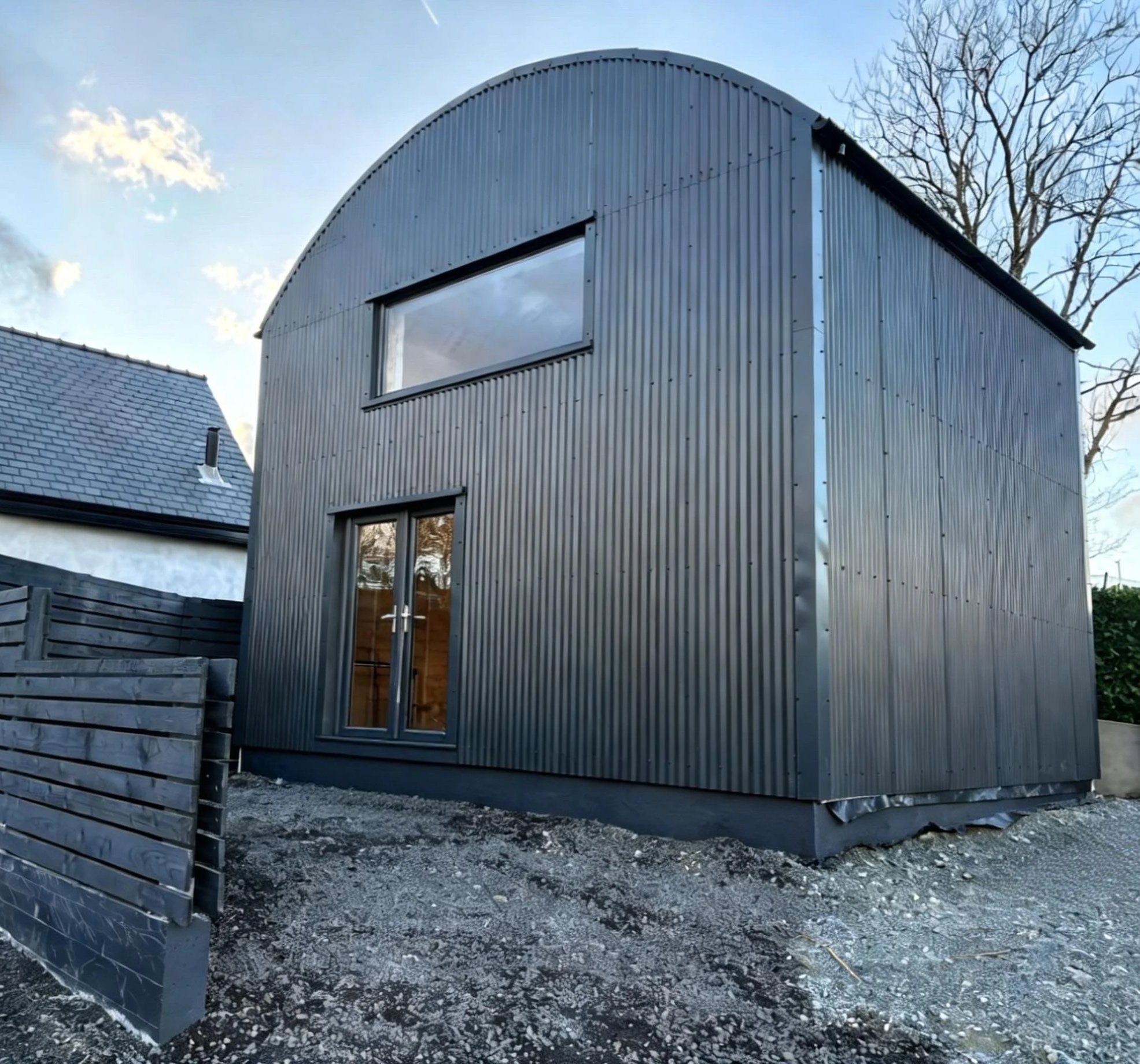 A modern metal building with curved roof and large horizontal window, next to a traditional house with a tiled roof and chimney. The image shows an outdoor scene with trees and a partially cloudy sky.