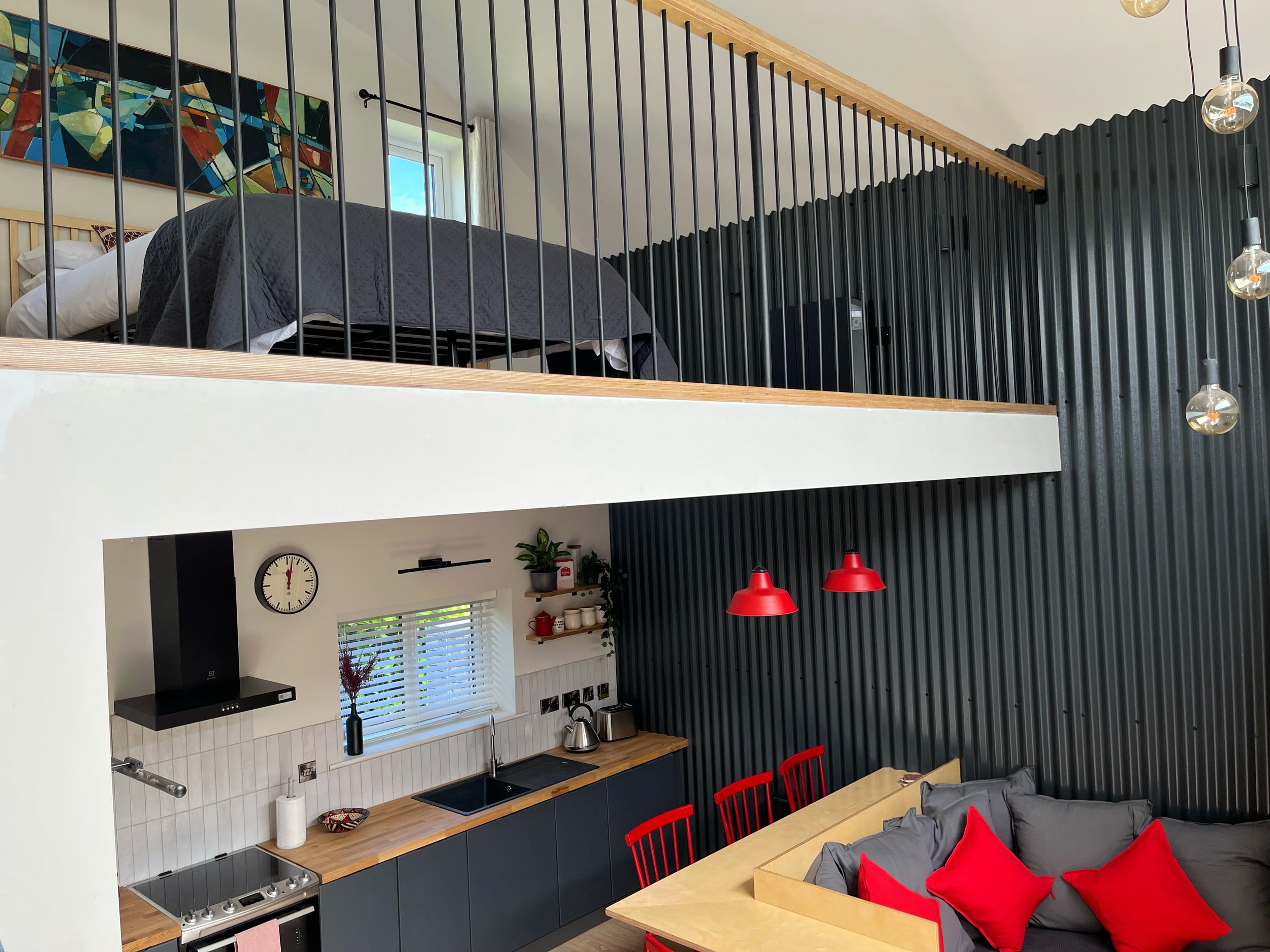 A modern kitchen and living area with a lofted bedroom. The kitchen has dark cabinets, a wooden countertop, a black sink, and a backsplash with white tiles. There are red chairs around a wooden dining table with gray cushions and red pillows. The wal