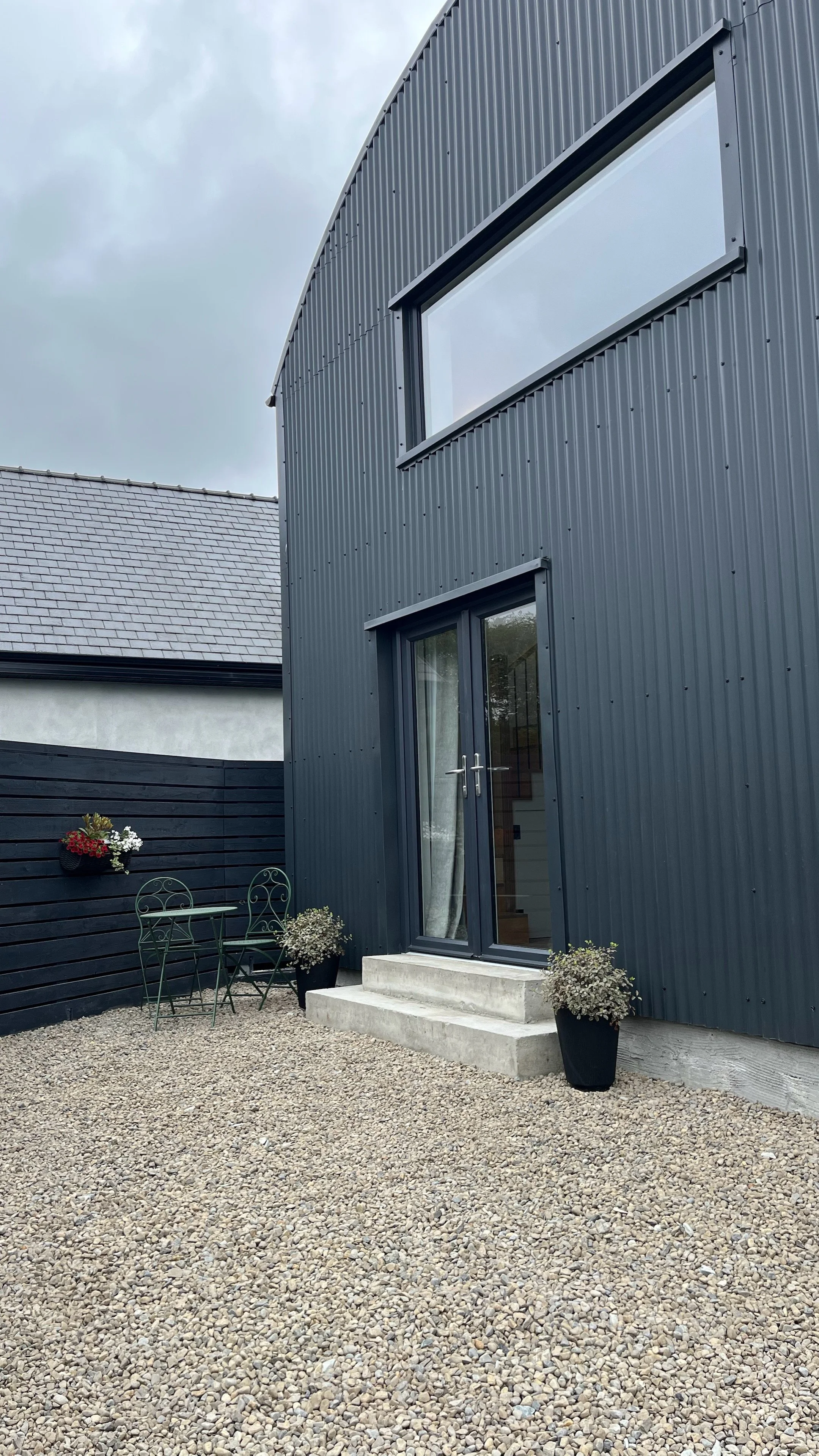 Modern gray house with black window frames, concrete steps at the entrance, and an outdoor seating area with a small table, chairs, and potted plants. A wooden fence with a flower arrangement is on the left side, and a gravel yard surrounds the house