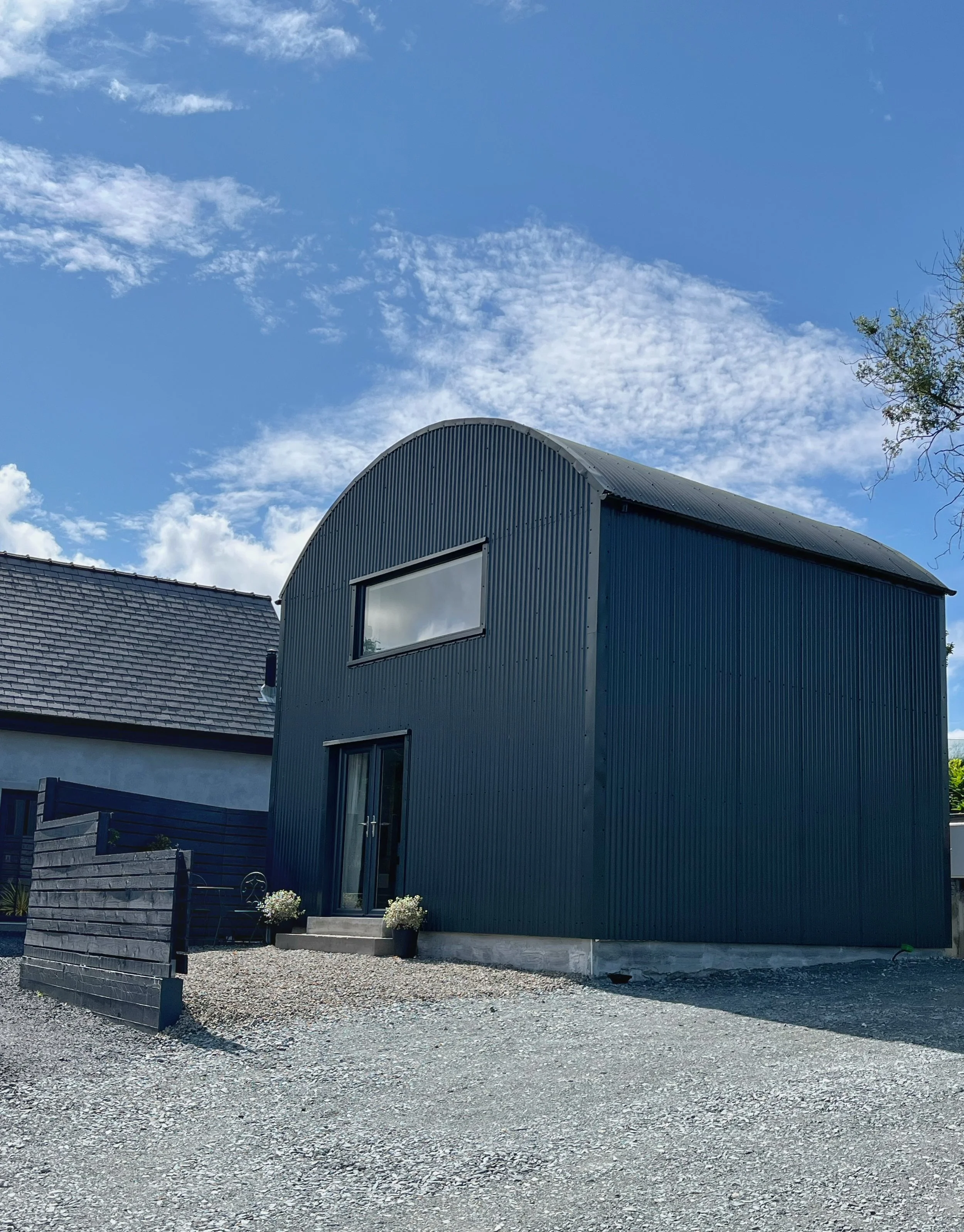 A modern blue building with a large window and a rounded roof, set against a partly cloudy sky. There are small flowers in pots near the entrance and a gravel ground in the foreground.