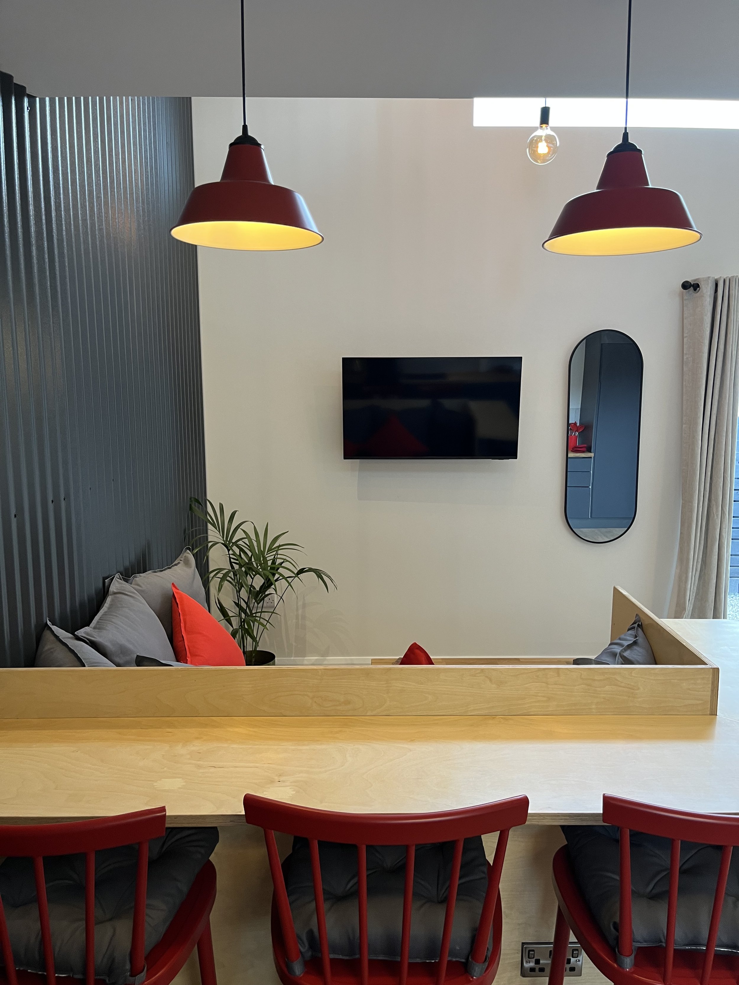 View of a modern living room with a black wall, wall-mounted flat screen TV, oval mirror, curtain, and a corner with a plant. The ceiling has two red pendant lights and a small exposed bulb. A person is sitting at a wooden table with red chairs.