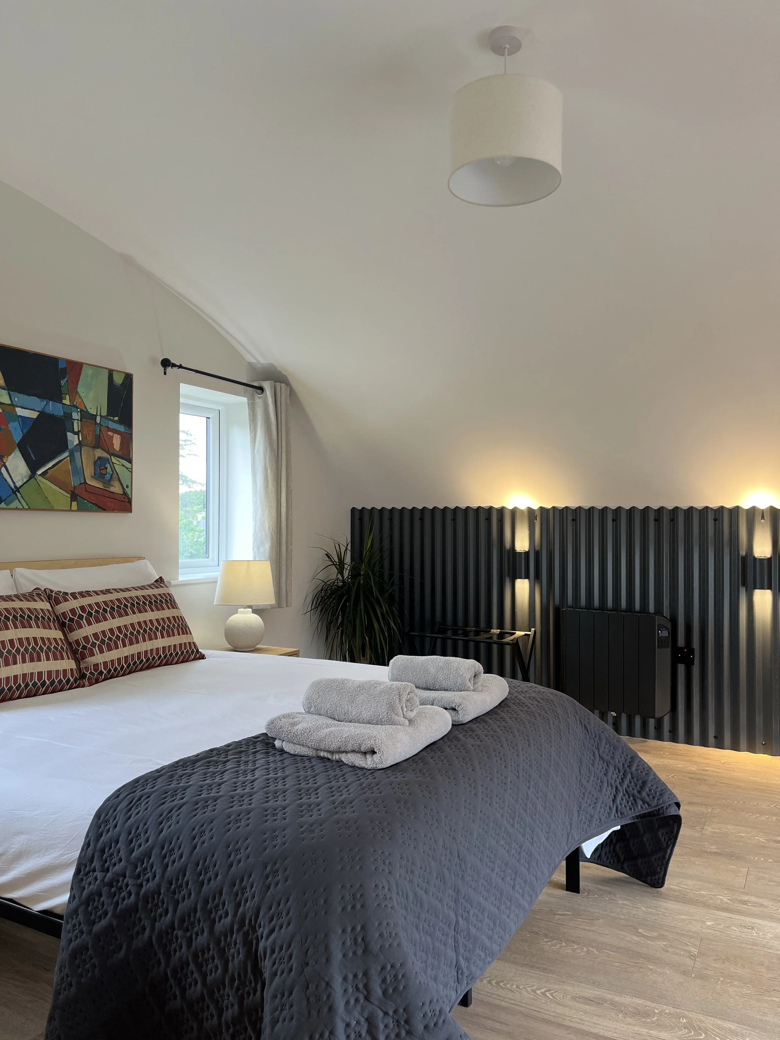 A bedroom with a bed covered in a dark gray quilt, topped with three folded white towels. The headboard is black and textured. A nightstand with a white lamp is beside the bed. There are patterned pillows with red and beige colors. A small window wit