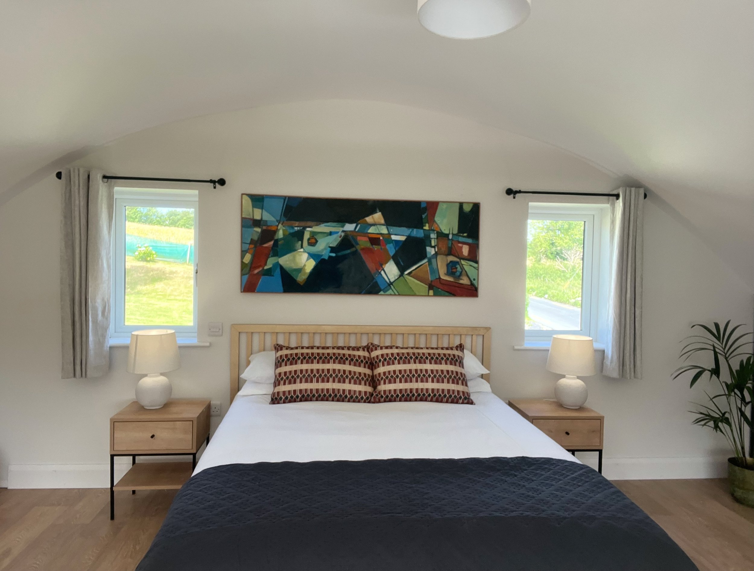 A cozy bedroom with a white bed, patterned pillows, and matching nightstands with lamps on each side. Two windows with white curtains and an abstract colorful painting above the bed, with trees visible outside through the windows.