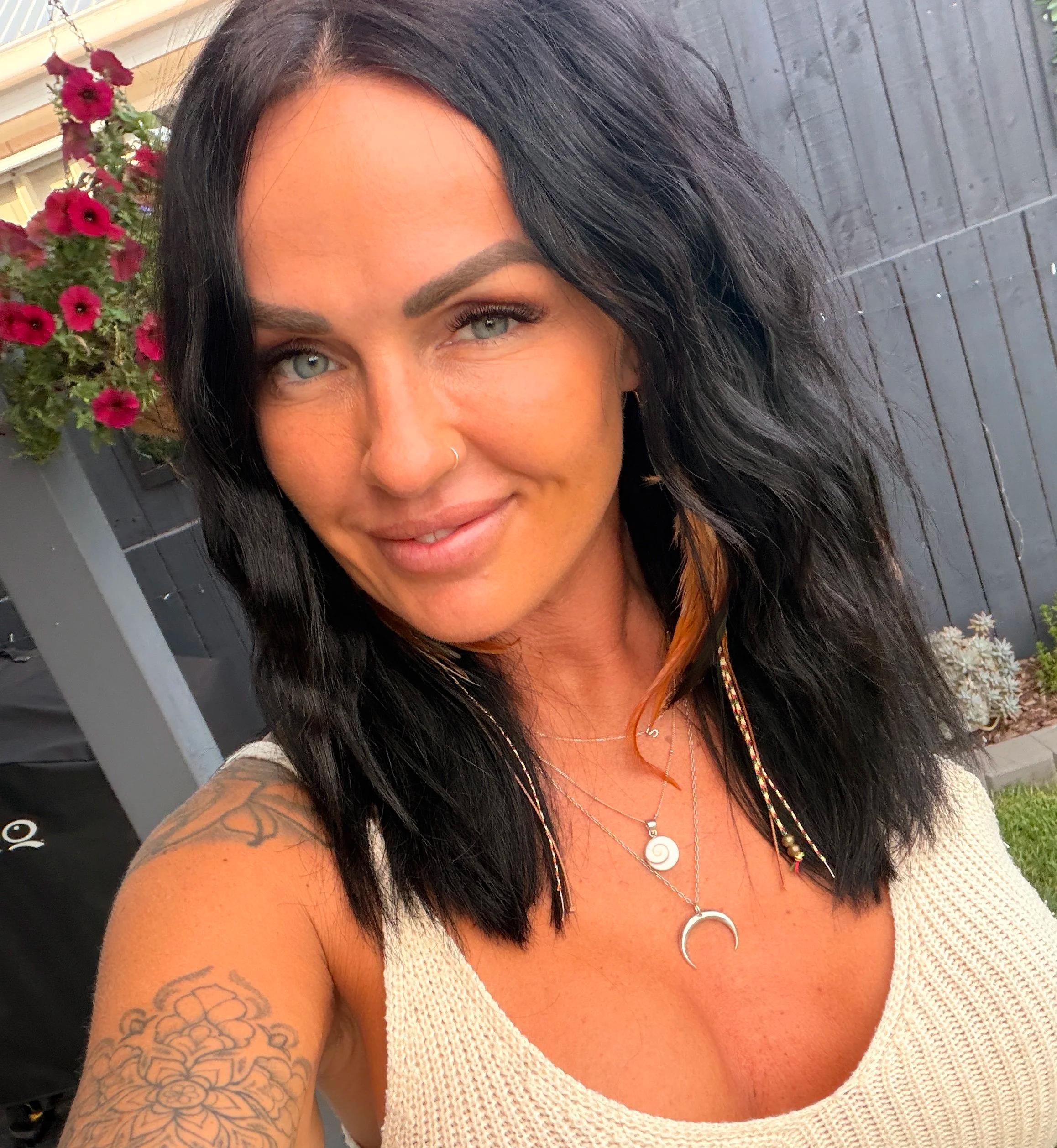 A woman with black wavy hair, green eyes, and tattoos on her shoulder, smiling outdoors with flowers and a gray wooden fence in the background.