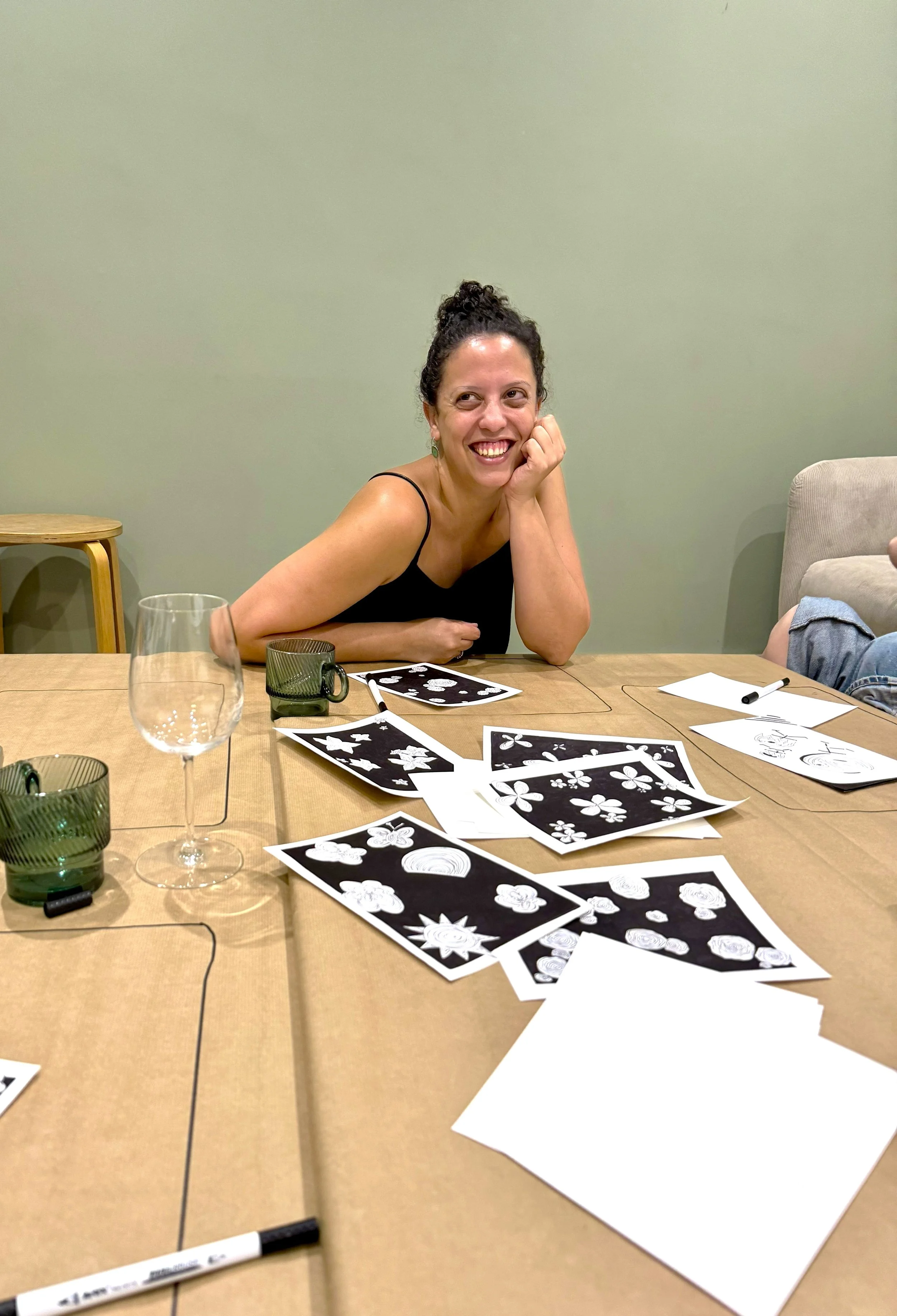 A woman with dark curly hair in a bun, wearing a black spaghetti strap top, is sitting at a table and smiling while resting her chin on her hand. The table has black and white illustrated cards spread out, some blank sheets of paper, markers, and glass cups. A wooden chair with arms is in the background against a plain green wall.