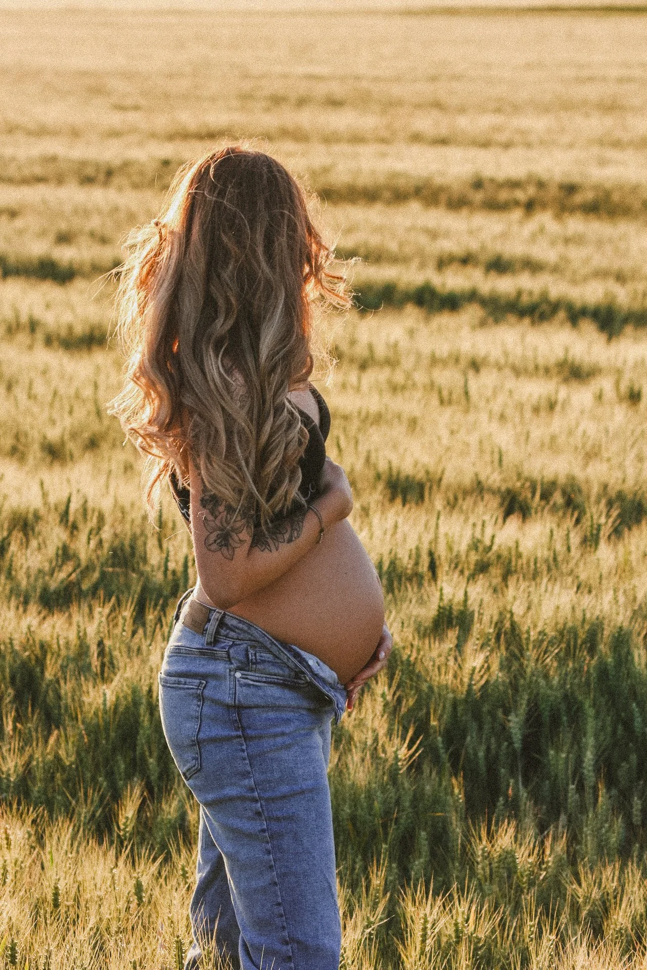 Schwangere Frau mit langen, lockigen Haaren steht in einem goldenen Getreidefeld bei Sonnenuntergang, die Hände auf den Bauch legend.