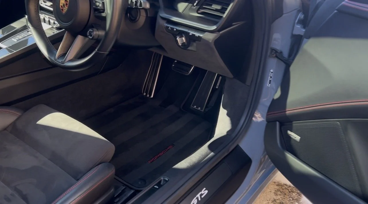 Interior of a Porsche sports car showing the driver's side footwell, pedals, steering wheel, and a portion of the door with a speaker.