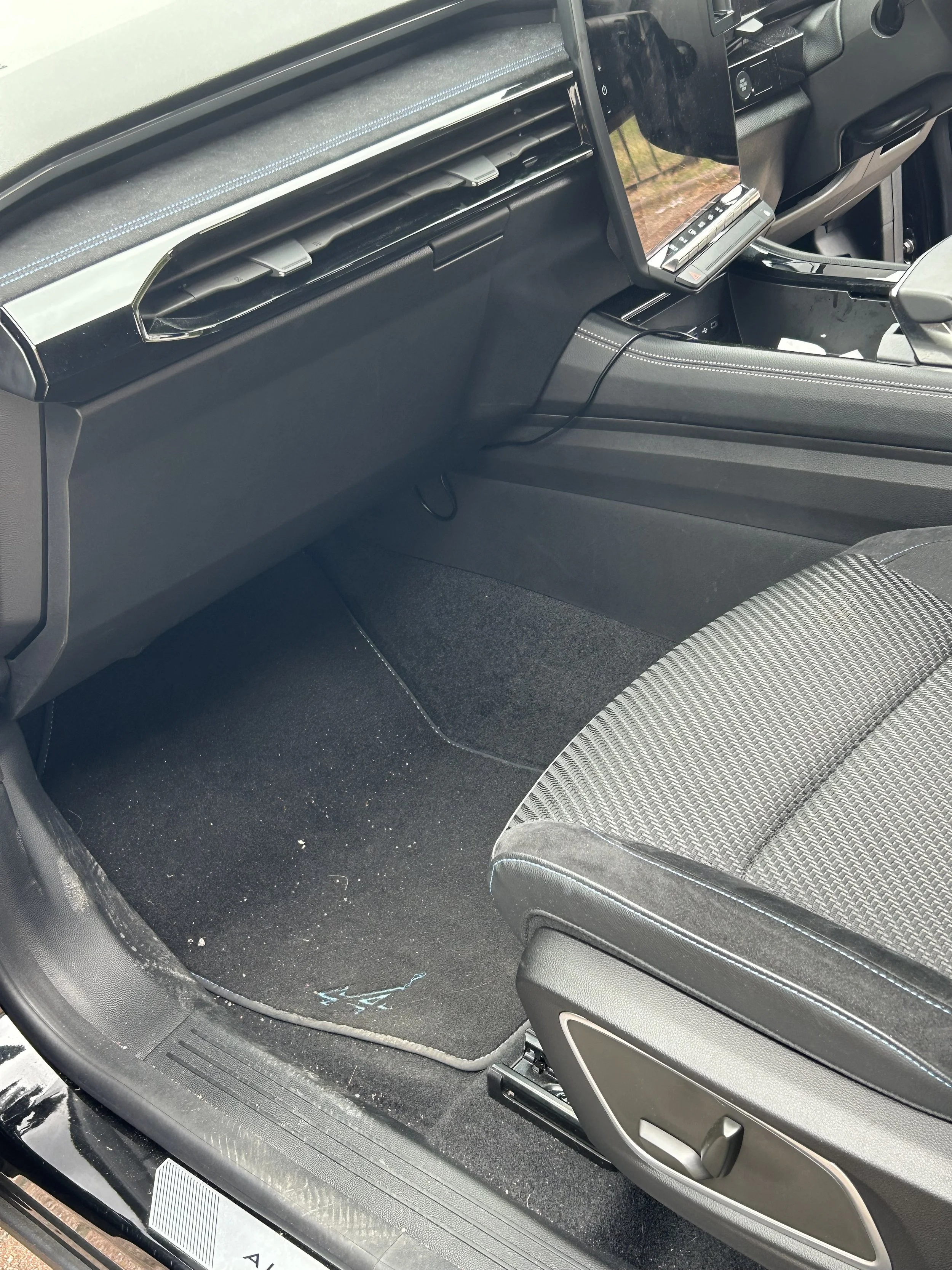 The interior of a vehicle showing the passenger side floor, with the door open, black carpet, and a patterned fabric seat.