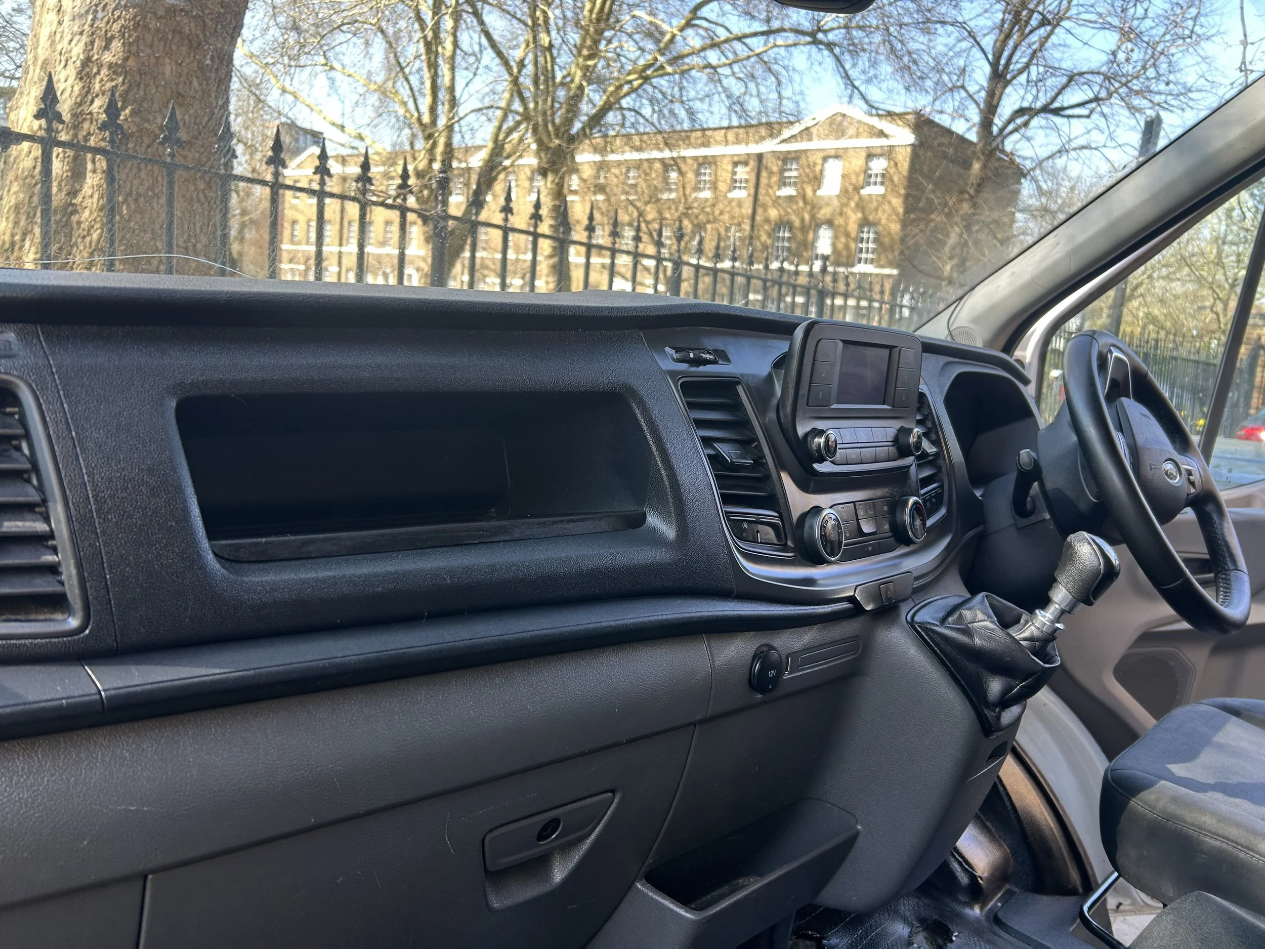 View of the interior of a vehicle dashboard and steering wheel with a large windshield showing a building and trees outside.