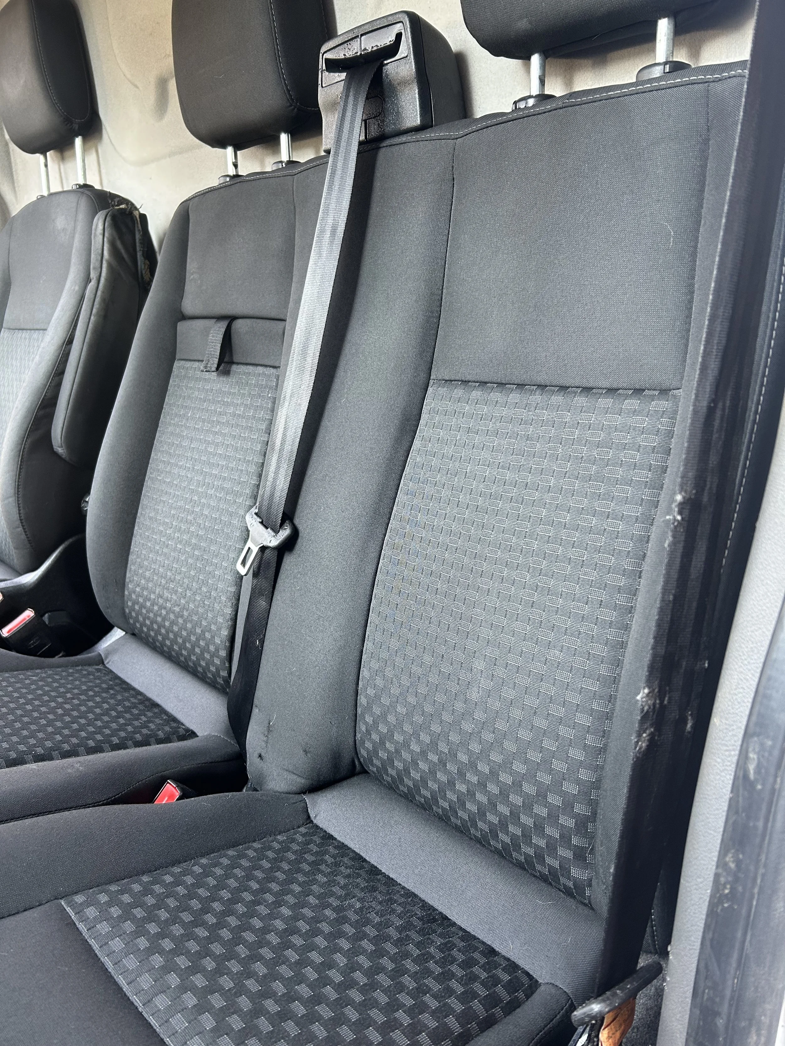The image shows the back seats of a vehicle with black and gray fabric upholstery, a fabric-covered floor, seat belts, and headrests.