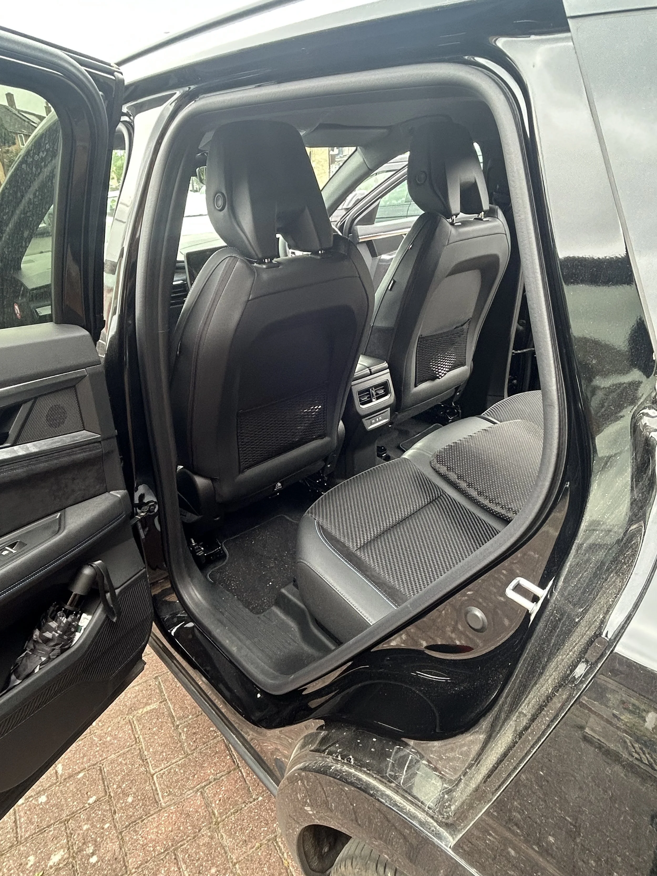 The interior of a black SUV with two visible front seats, controls and a digital display, parked on a brick driveway.