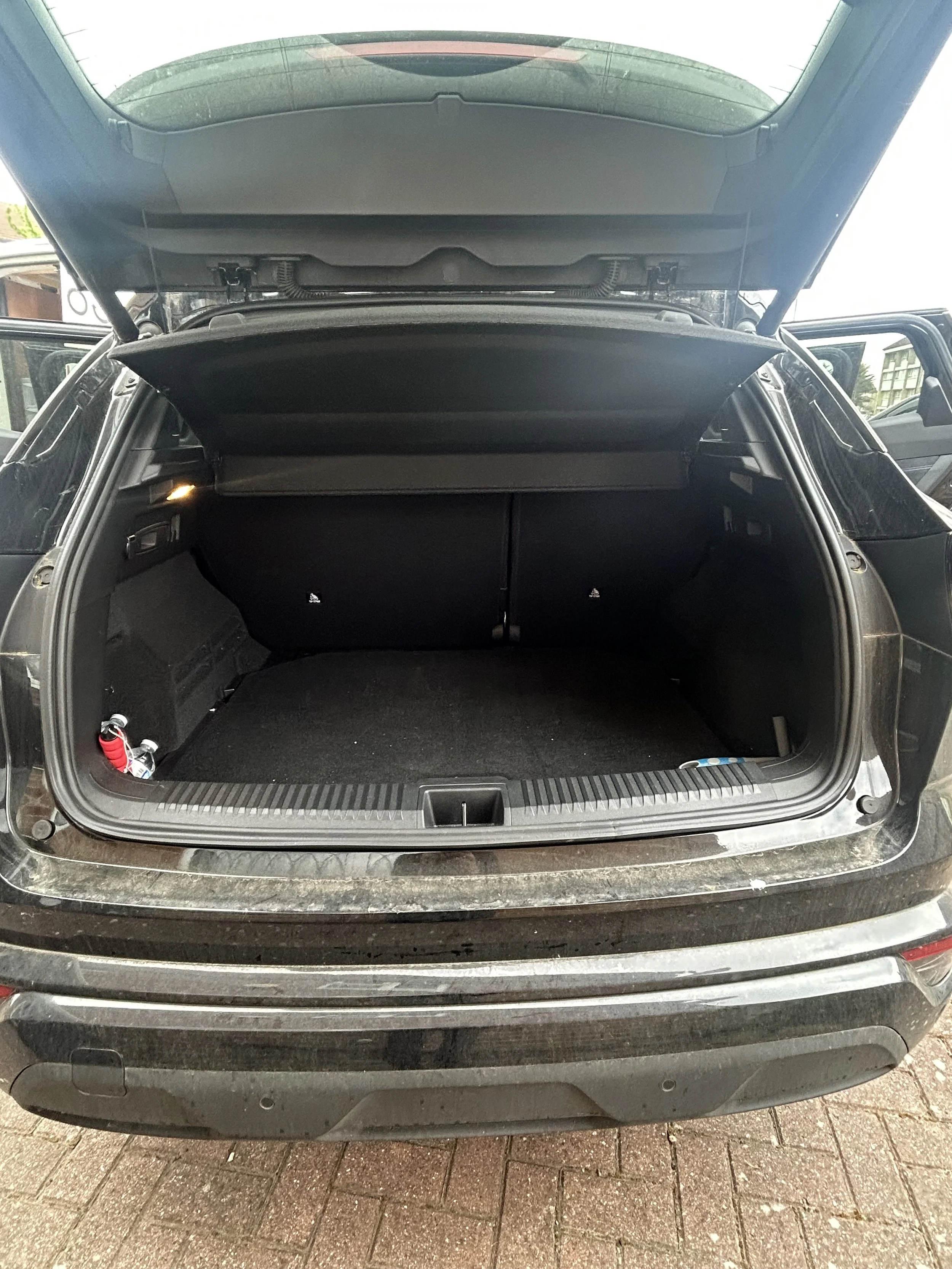 The trunk of a black SUV open, showing an empty cargo space with a retractable cover. The vehicle is parked on a brick driveway.