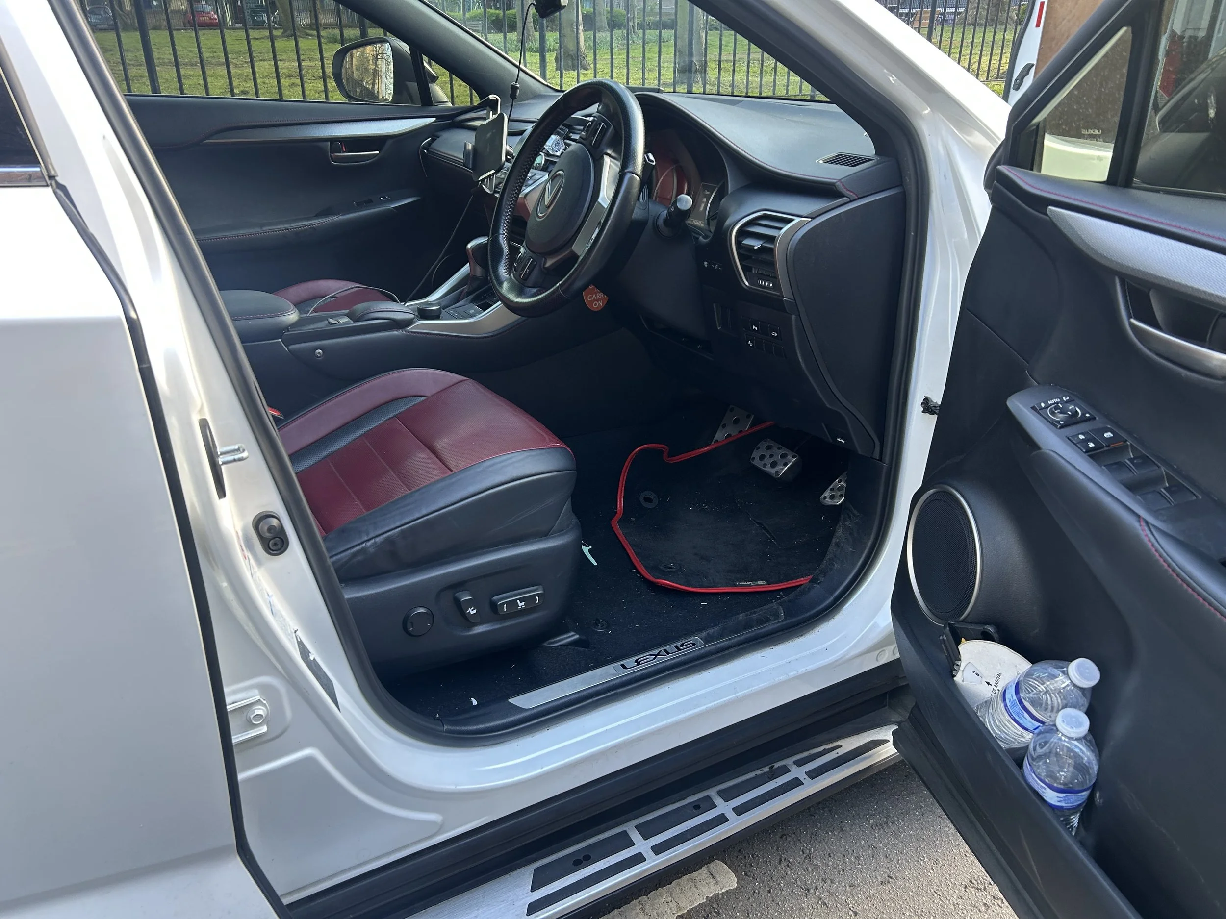 The interior of a white Lexus SUV showing the driver's seat, steering wheel, dashboard, center console, and part of the passenger seat with red and black upholstery. There are water bottles on the ground outside the open door.