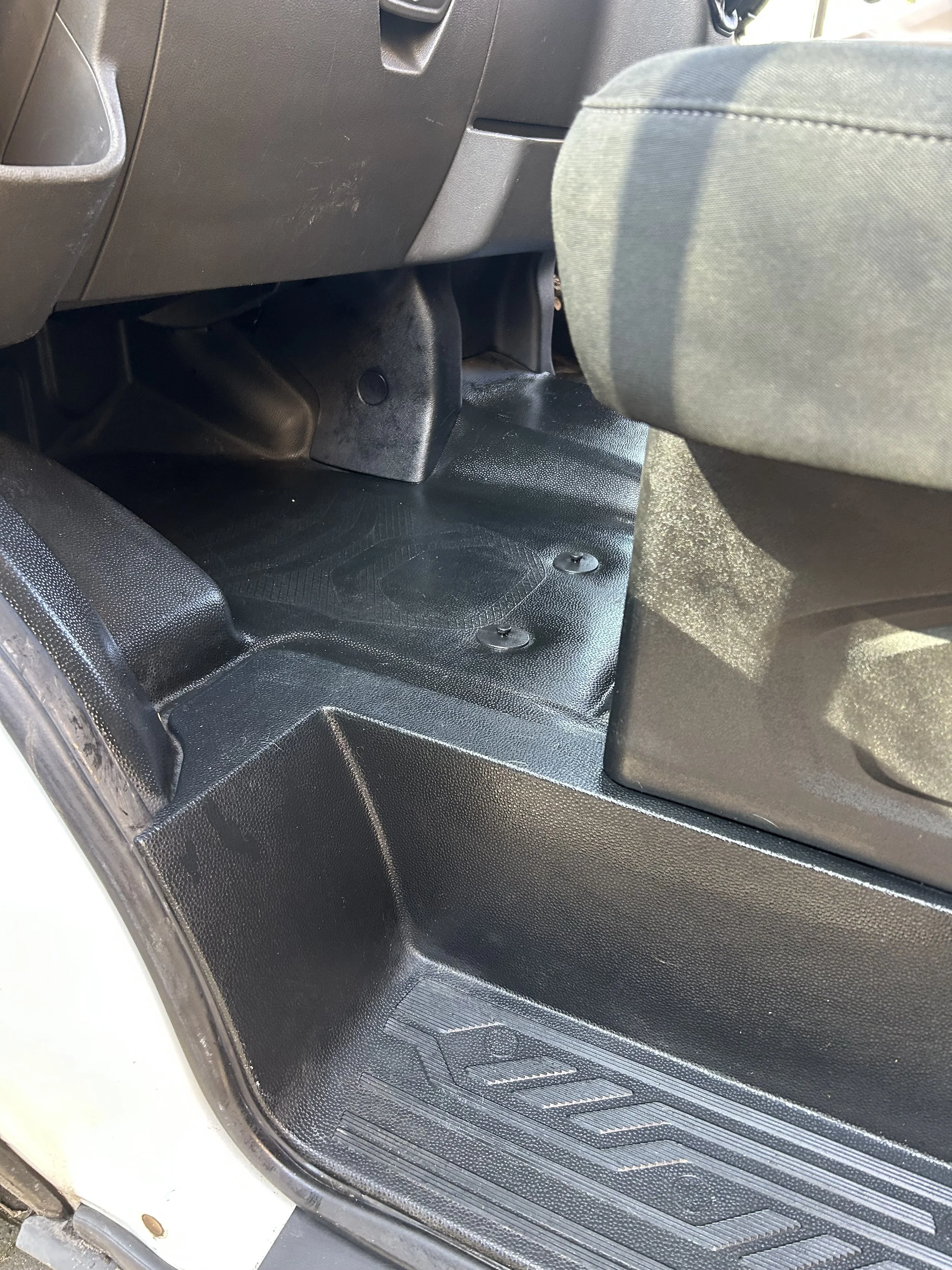 The image shows the interior of a vehicle focusing on the control panel behind the front seats. It includes a black, textured floor mat, the back of a passenger seat with grey fabric upholstery, and part of the vehicle's dashboard and side panels.