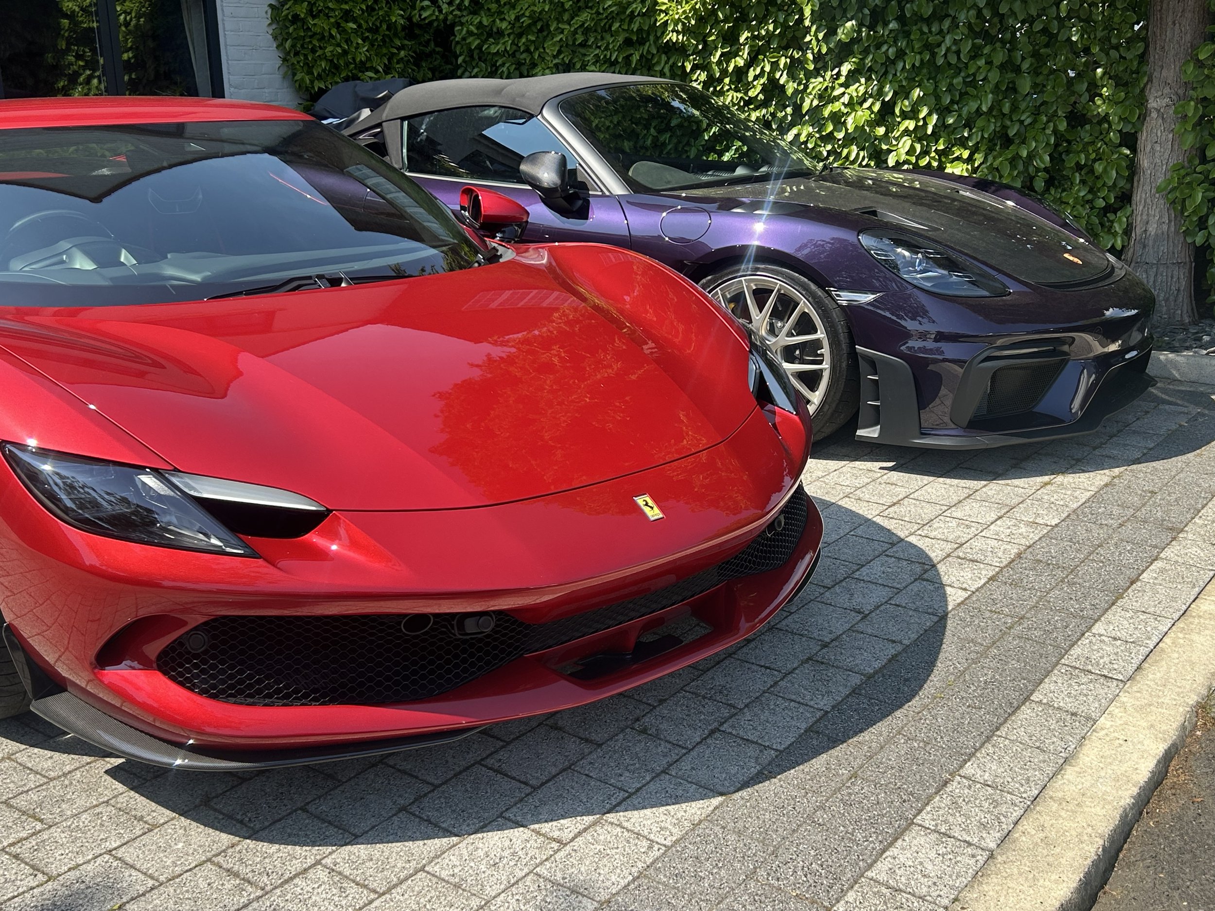 Red Ferrari sports car parked on a brick driveway, with a purple convertible car and greenery in the background.