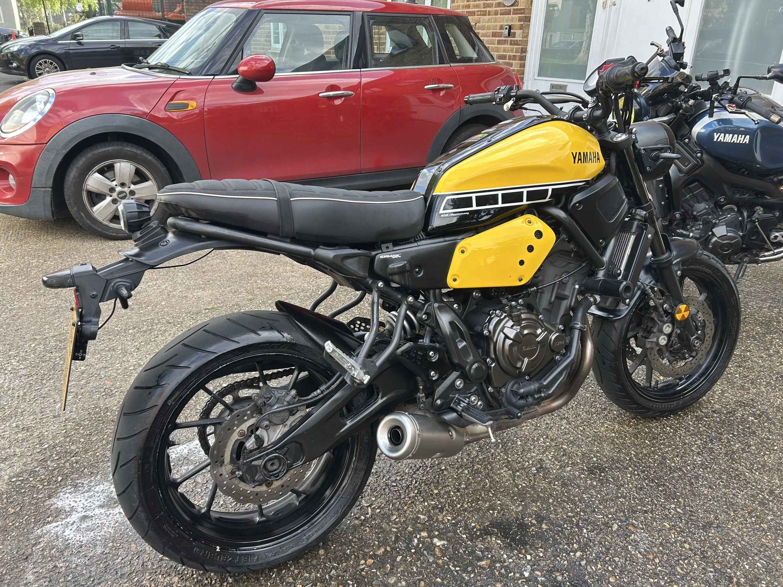 A red and black motorcycle with yellow accents parked in front of a red brick building, with a yellow car and a blue Yamaha motorcycle in the background.