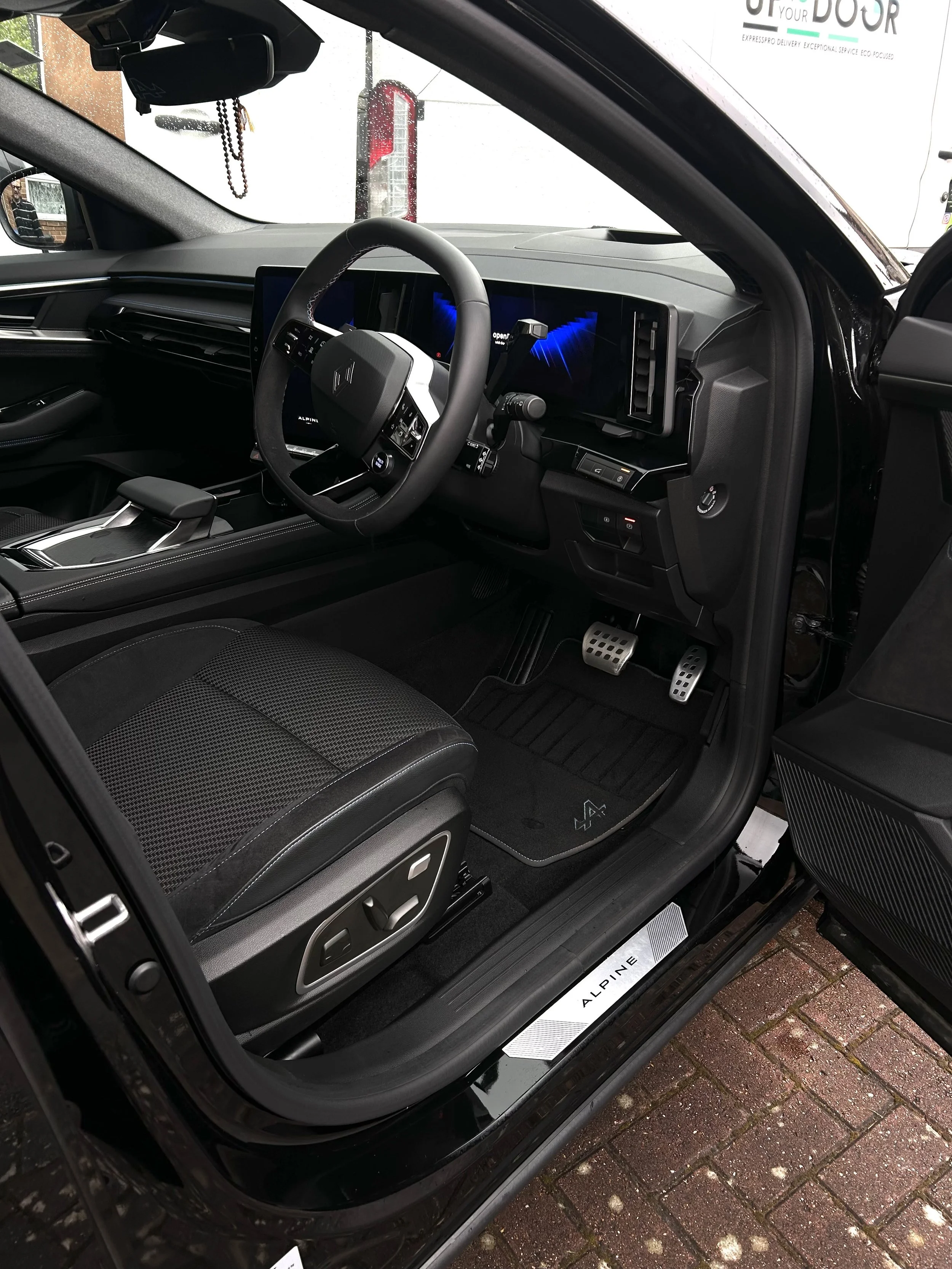 Interior view of a modern car dashboard, steering wheel, dashboard display, center console, adjustable seat, pedal area, and floor mat with the brand name 'Alpine' visible on the door sill.