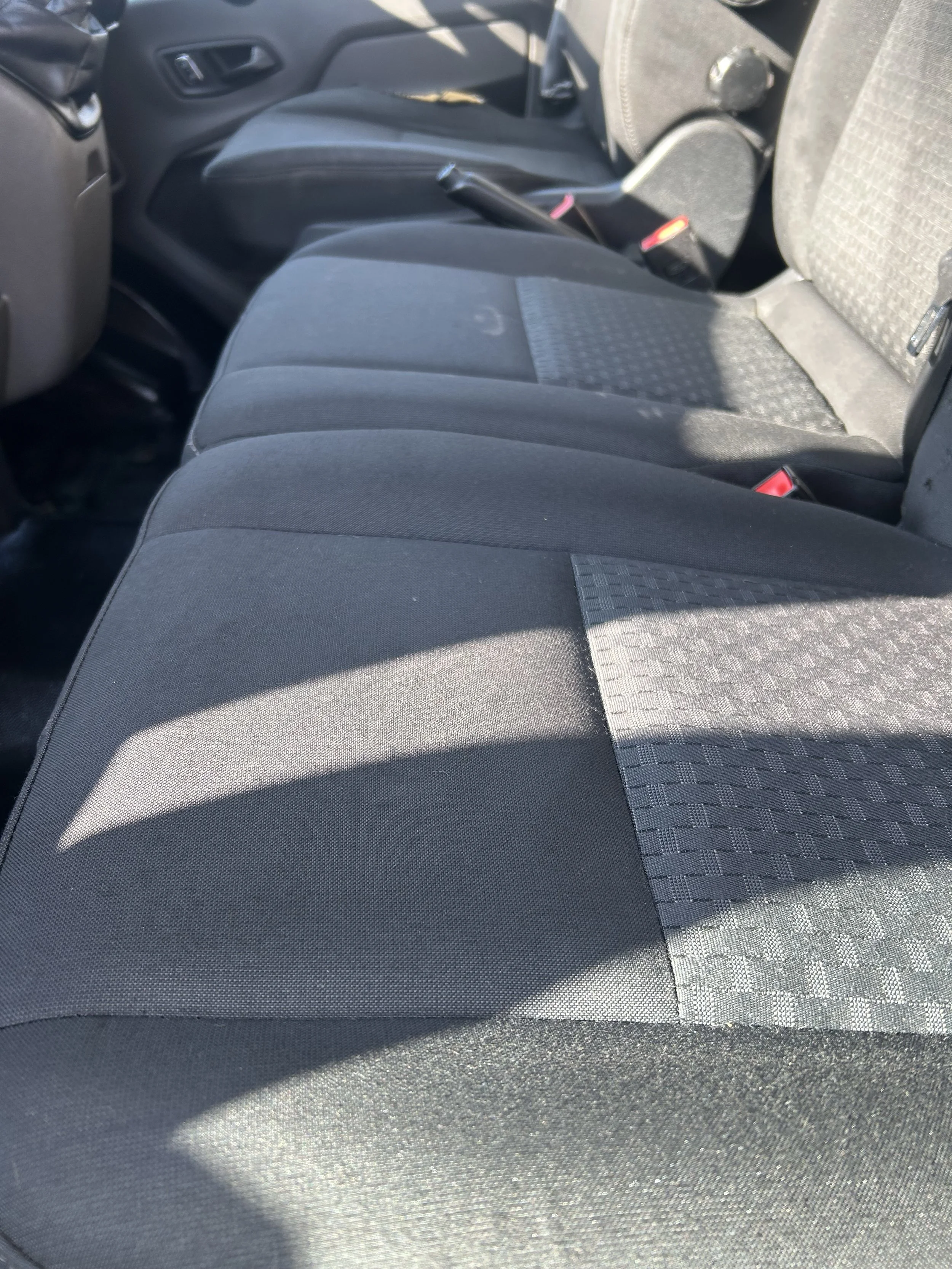 Inside a vehicle showing the front passenger seat and part of the driver's seat with fabric upholstery and safety seatbelt buckles.