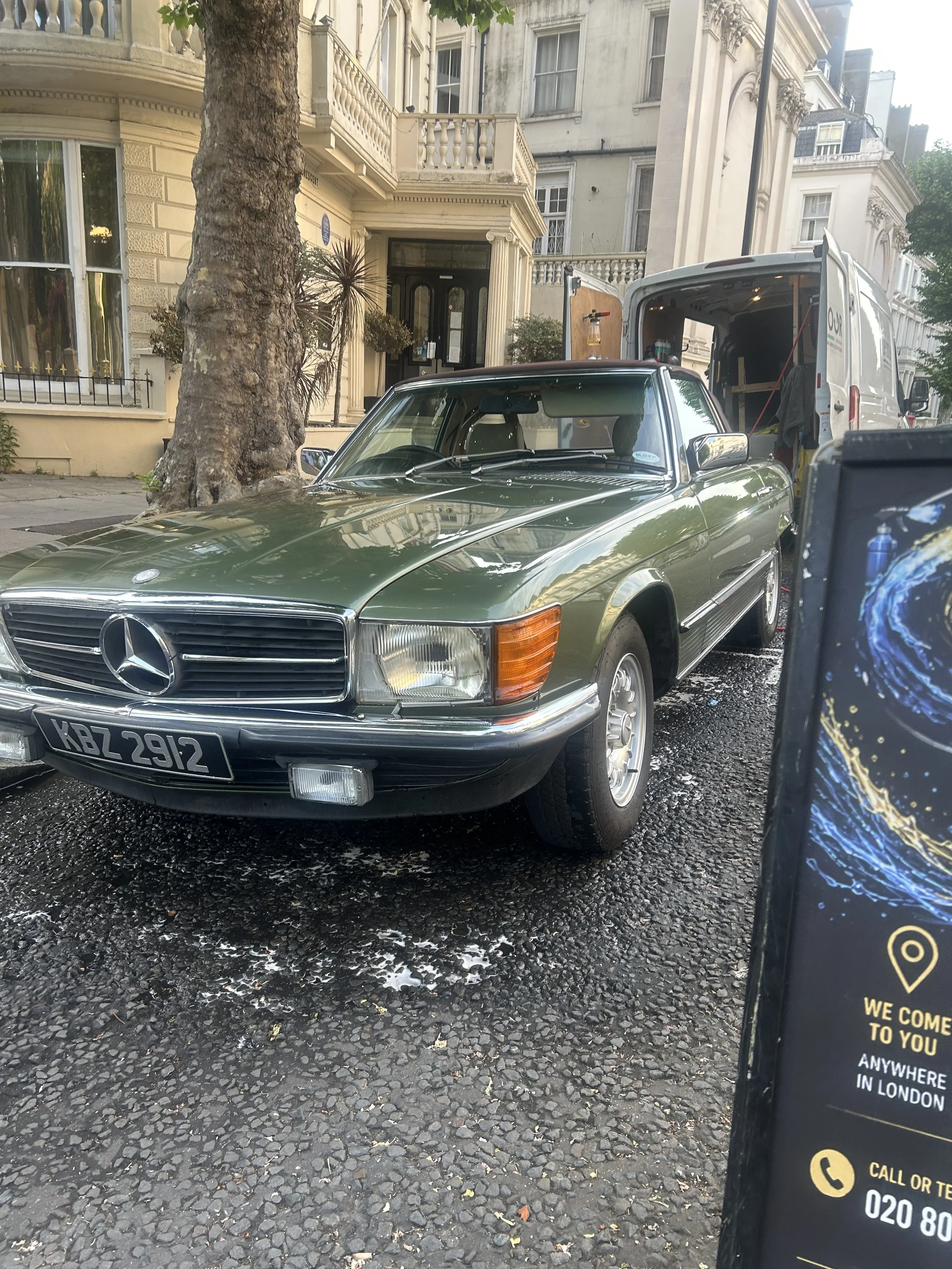 A classic green Mercedes-Benz car parked on a wet street in front of a large, ornate residential building with a tree in front. There is a partially visible delivery van behind the car and a phone screen in the bottom right corner showing a map and c