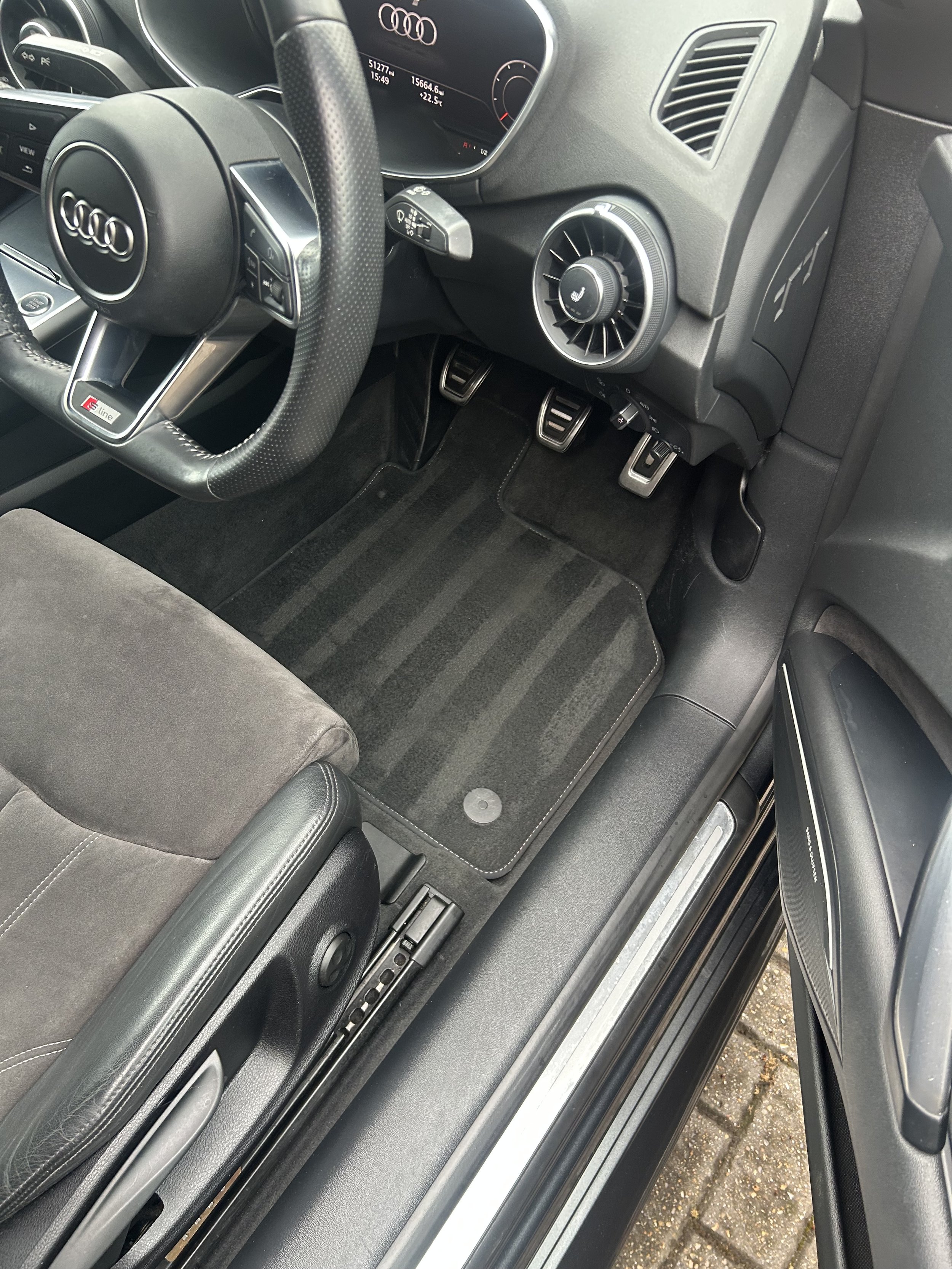 The image shows the interior of an Audi vehicle, focusing on the driver's side. It includes an Audi steering wheel with an S line badge, a digital dashboard display with information, and part of the center console with an air vent and control buttons