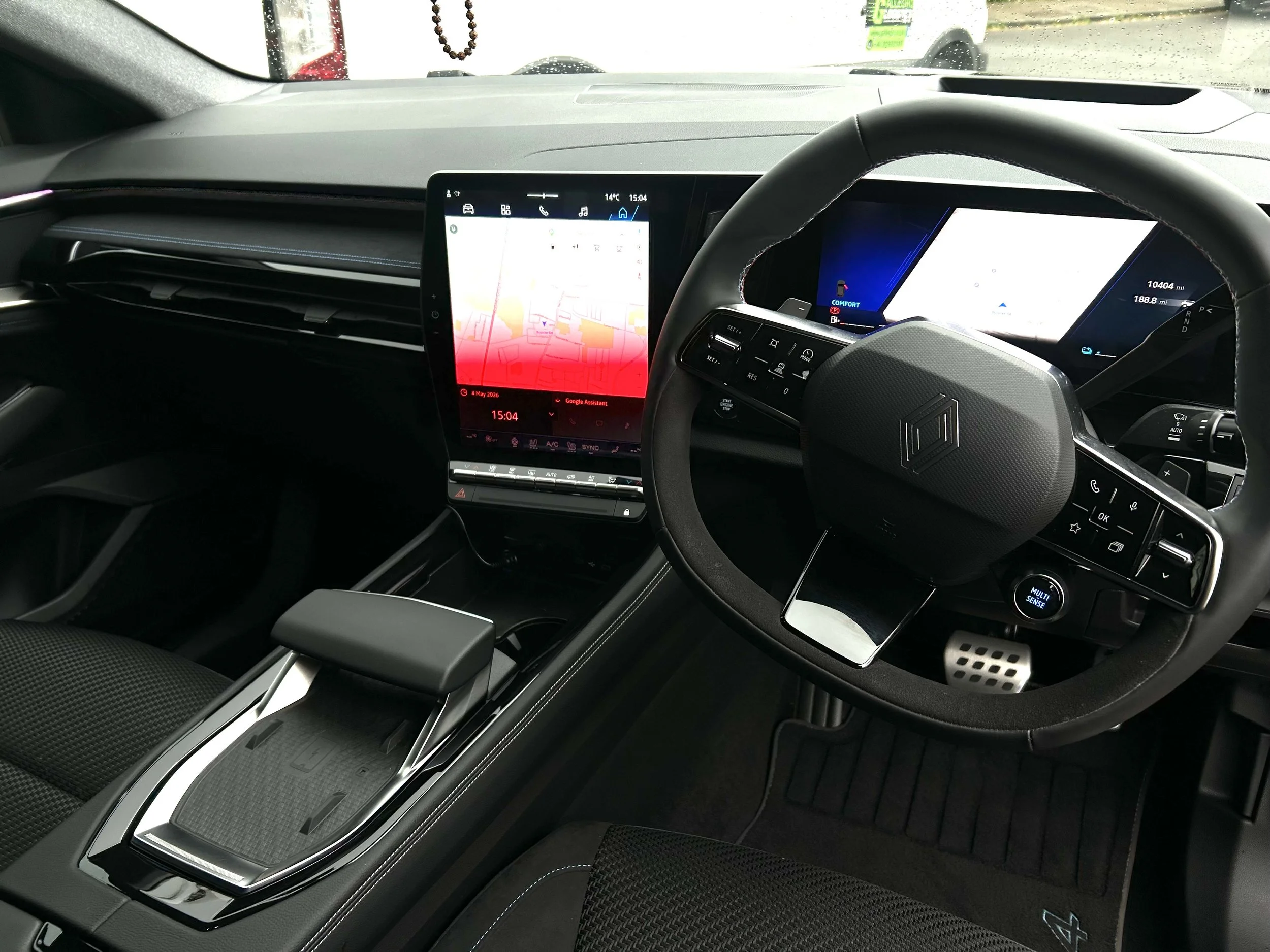 Interior of a modern vehicle showing a dashboard with multiple digital screens, steering wheel, and center console with controls, with raindrops visible on the windshield.