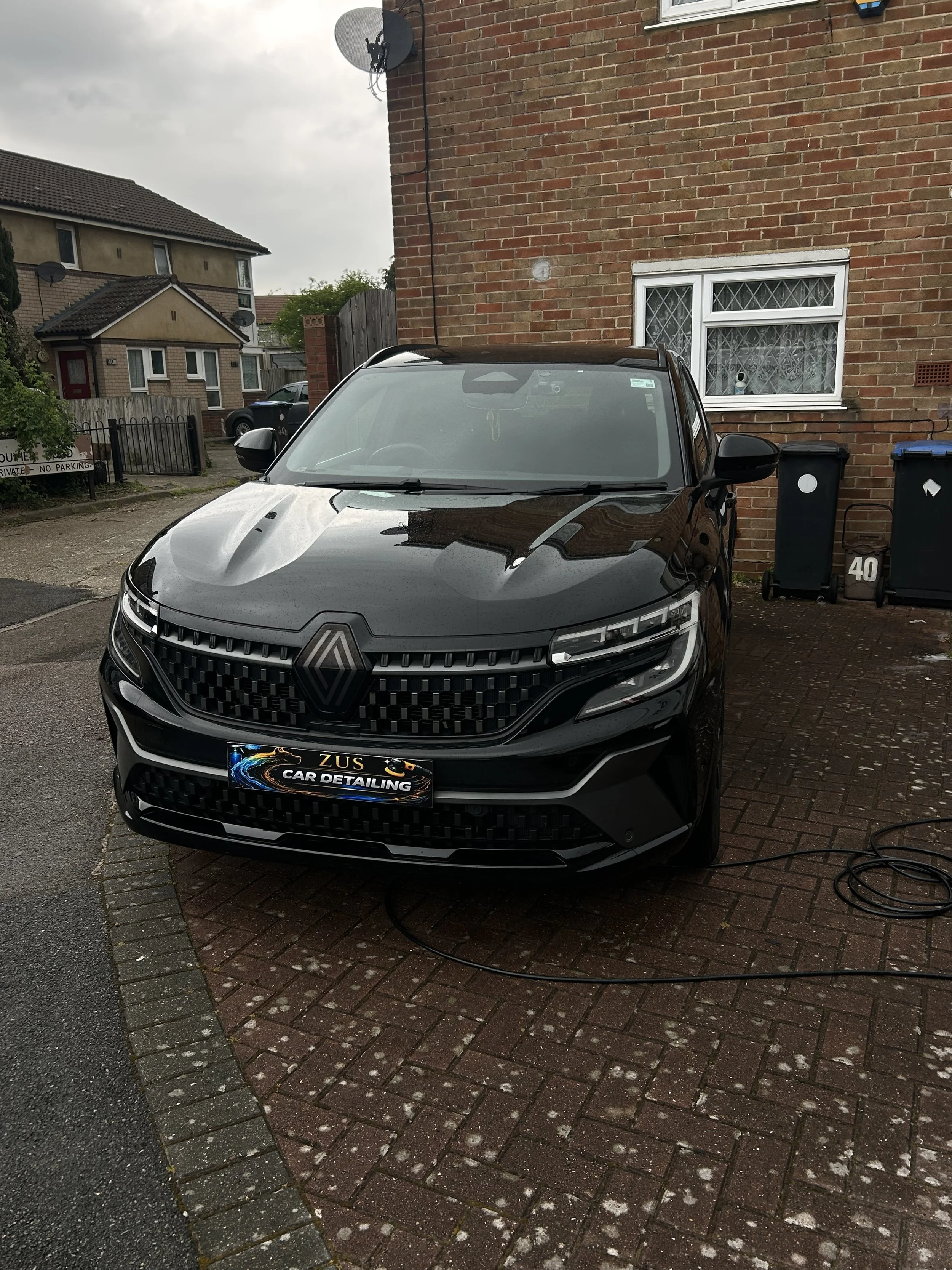 A black Renault car parked on a brick driveway outside a house with a brick exterior. The front of the car faces the camera, and it appears to be a recent model. The license plate reads 'ZUS CAR DETAILING'. The car is connected to a power cable, like