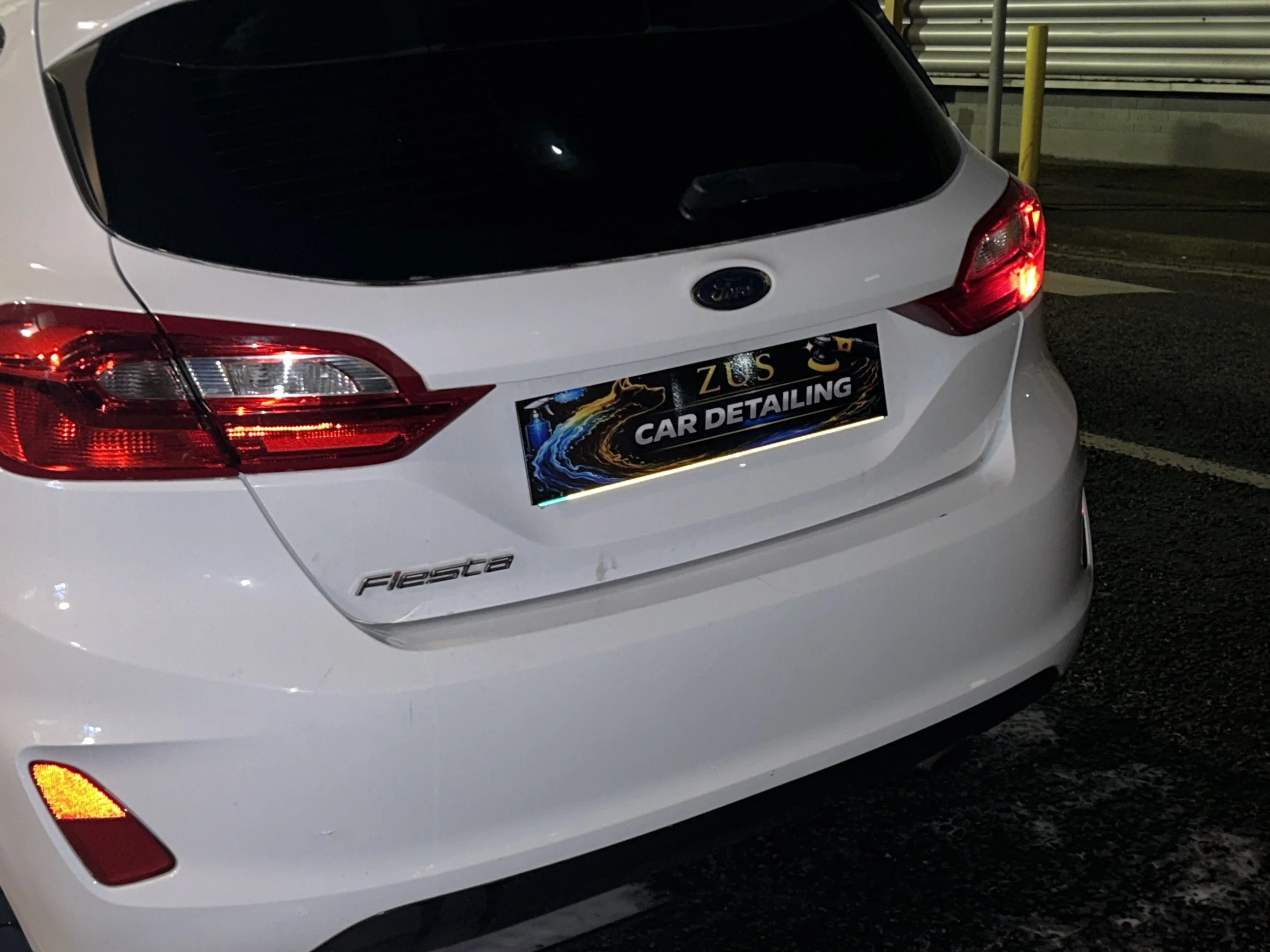 Rear view of a white Ford Fiesta with a car detailing advertisement on the license plate, parked in a dimly lit parking lot at night.