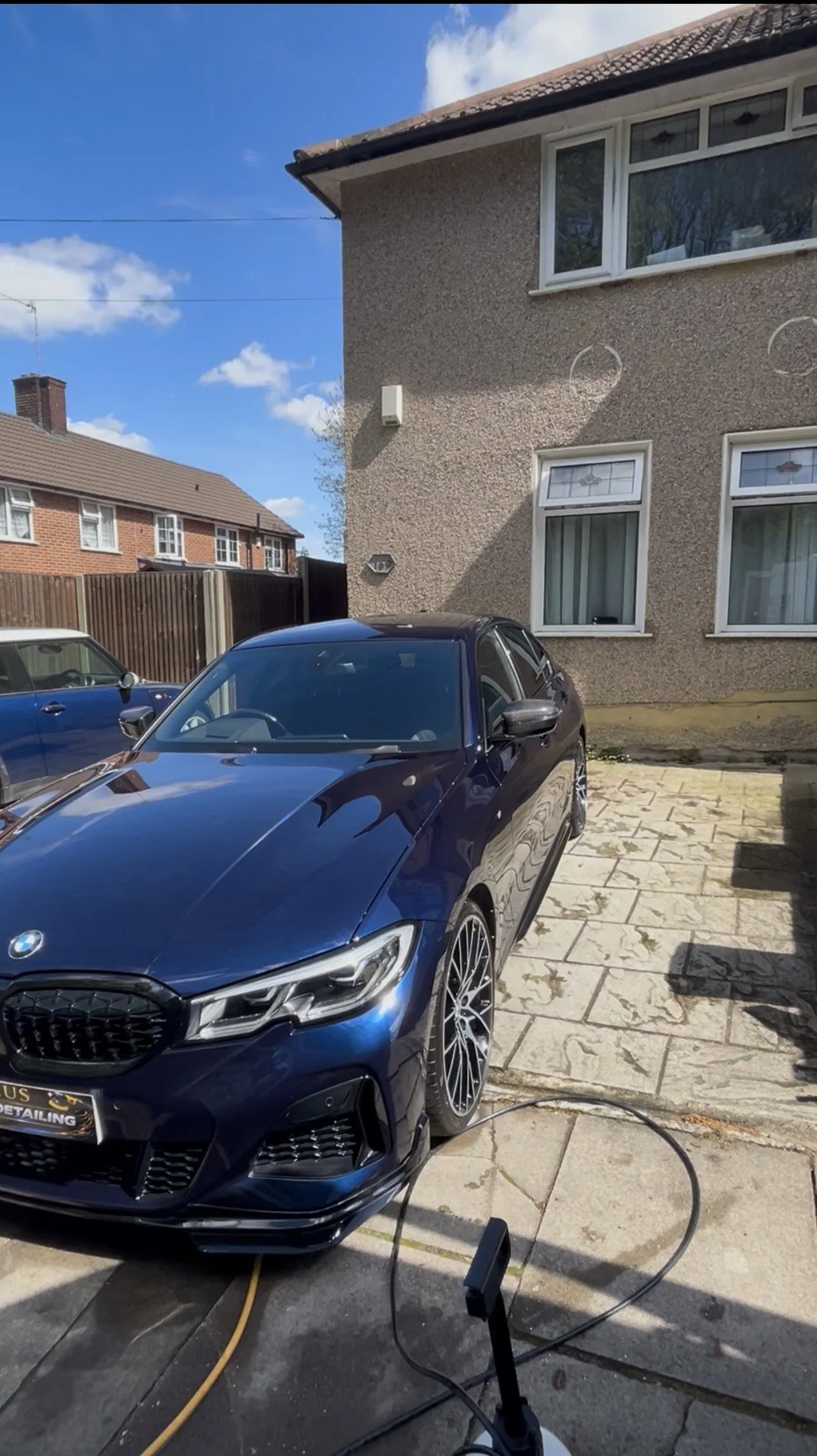 A black BMW car parked outdoors on a paved driveway in front of a beige house with white-framed windows. The driveway is being cleaned with a pressure washer, with a black hose and a water spray nozzle visible on the ground. The house has a textured 