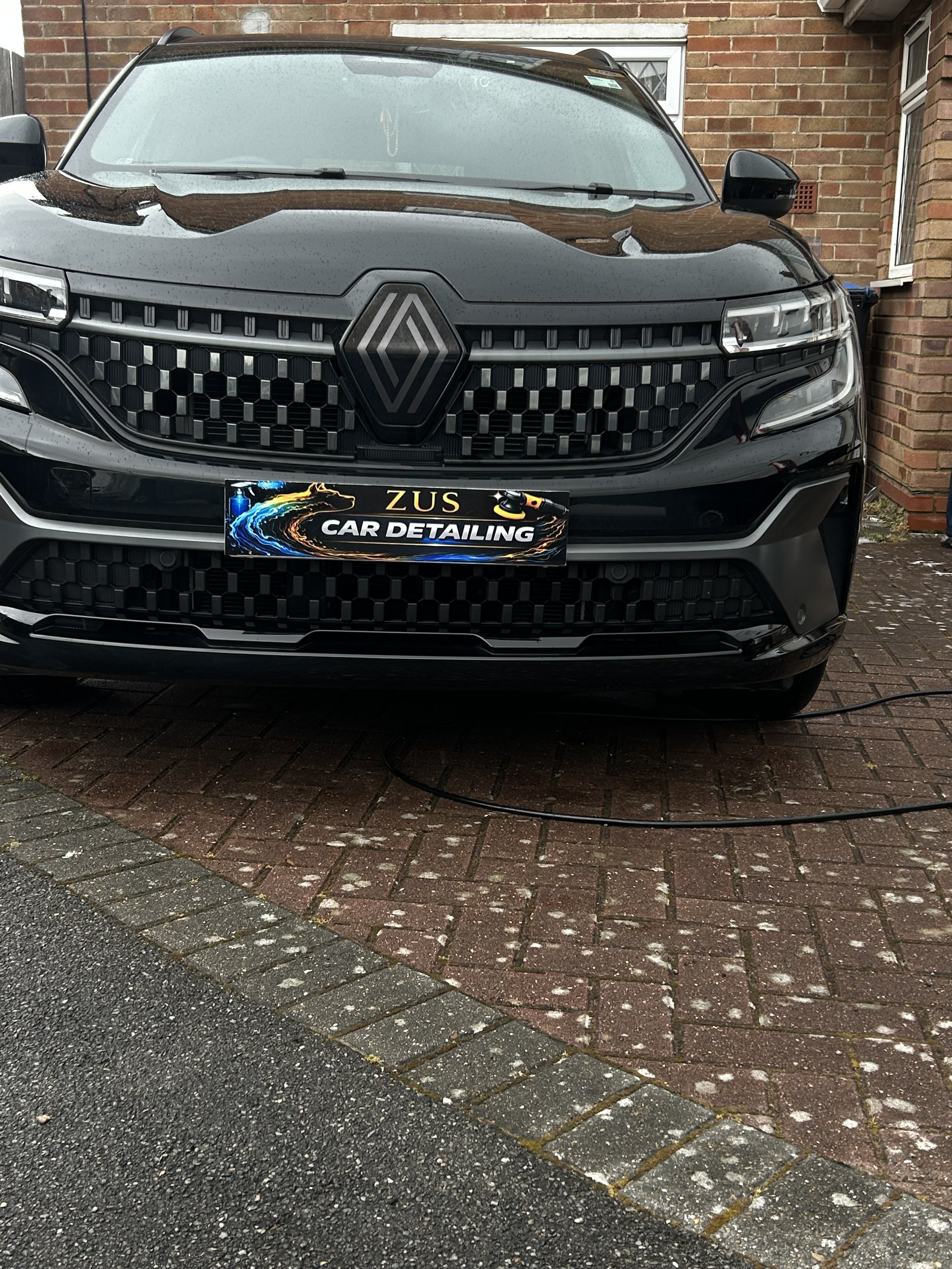 Black Renault car parked on a brick driveway outside a brick house with a small window. The car is wet with rain droplets, and a cleaning sign is on the front license plate. The driveway has a wet asphalt section and bricks.