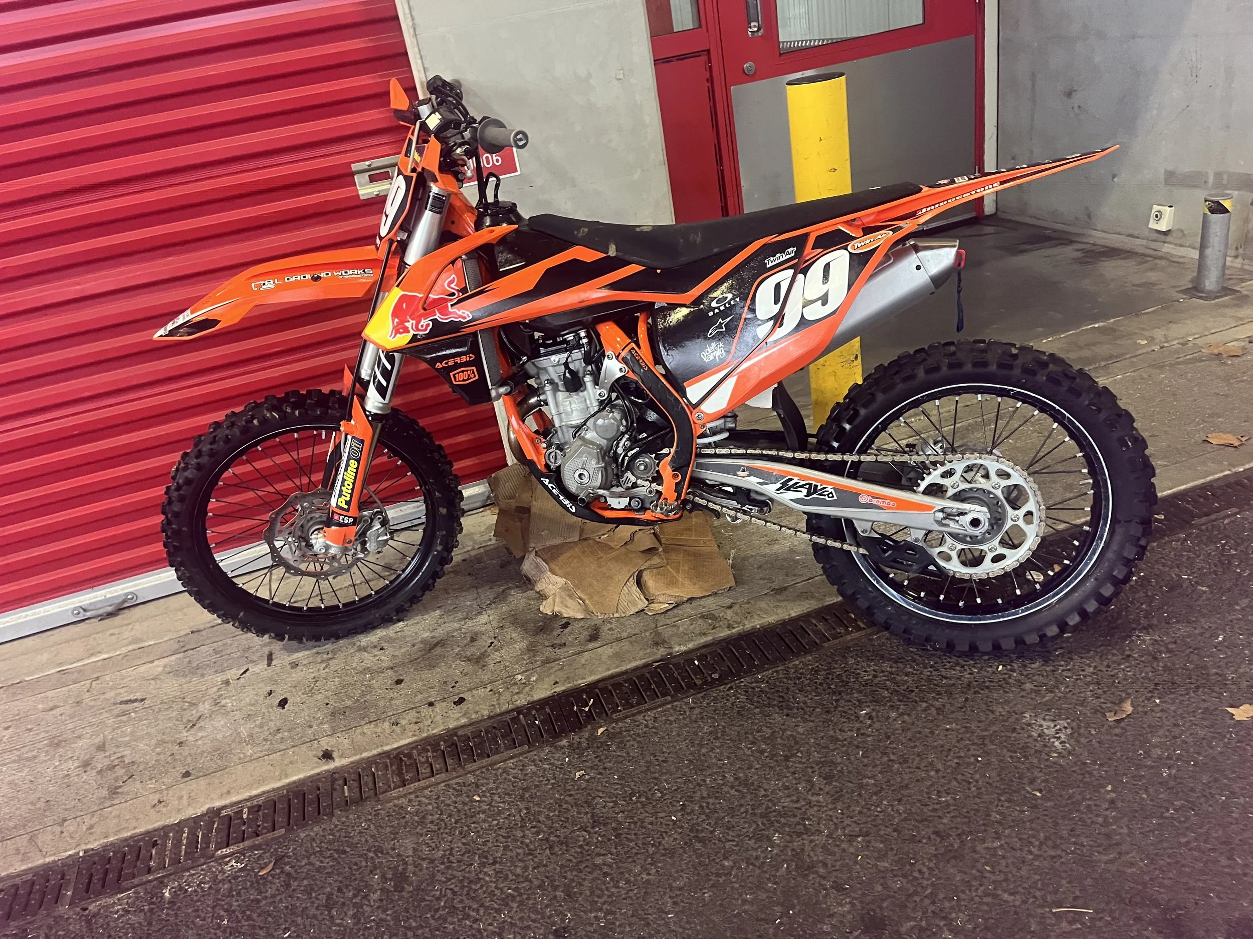 Orange and black dirt bike with racing number 99 against red and gray building wall.