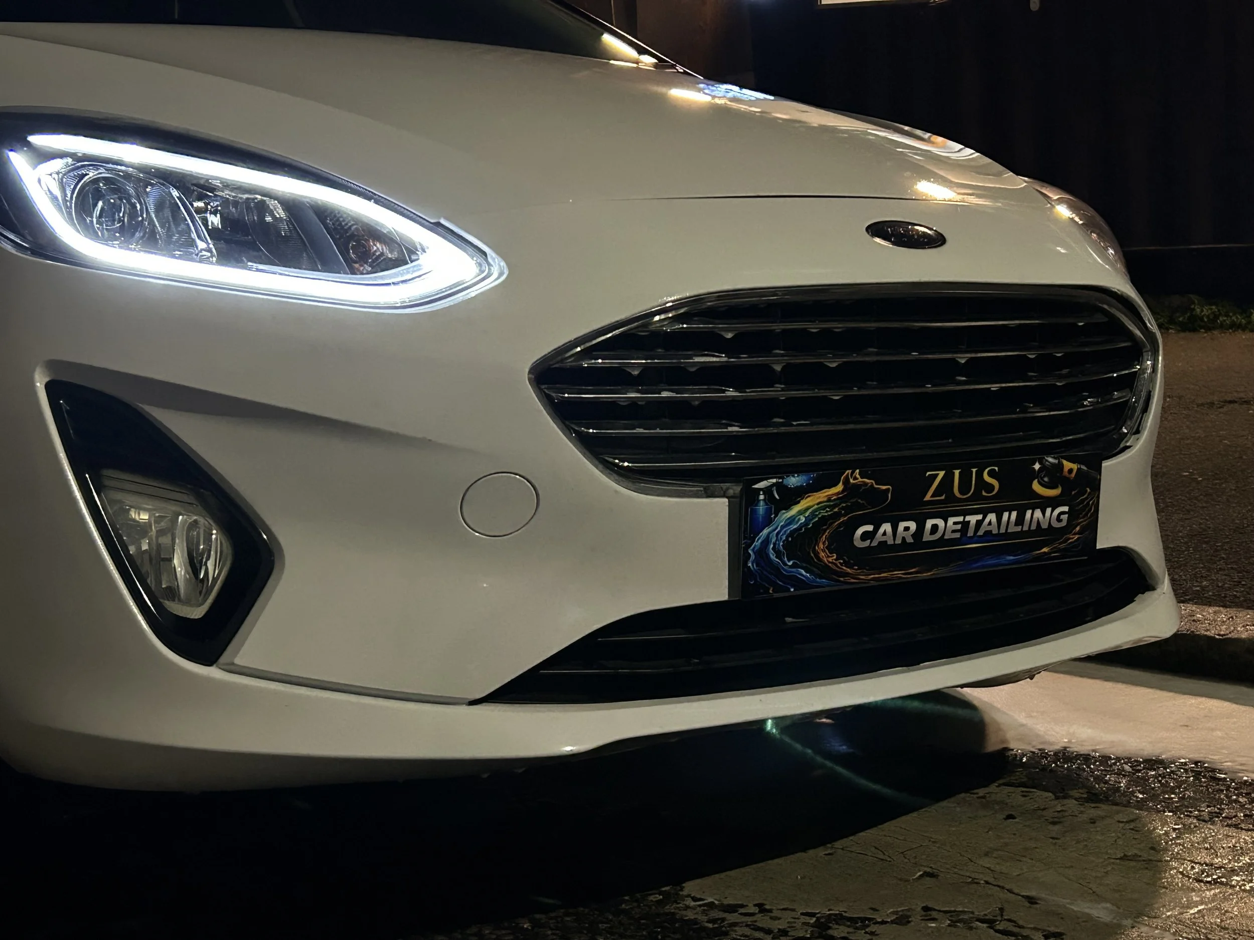 Front of a white modern car with headlights on, parked on a wet street at night. The car has a black license plate with 'ZUS CAR DETAILING' written on it.