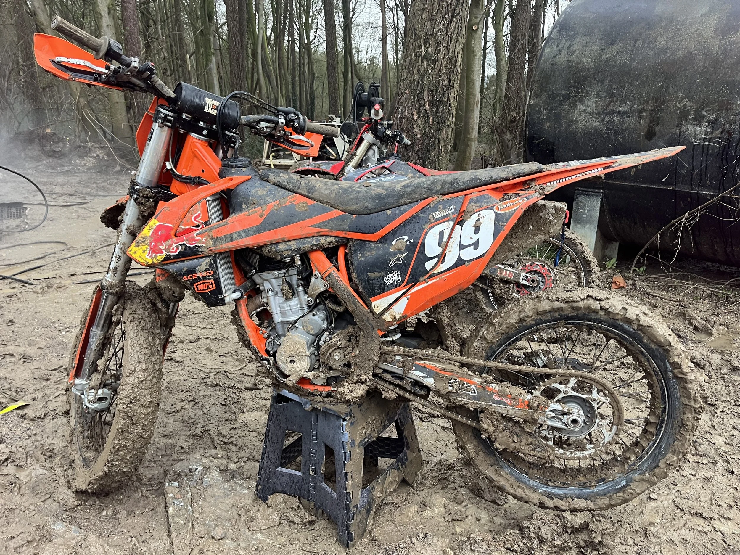 A damaged orange dirt bike with mud on the tires and body, leaning against a black stand in a muddy forest setting, with trees and a large black trailer in the background.