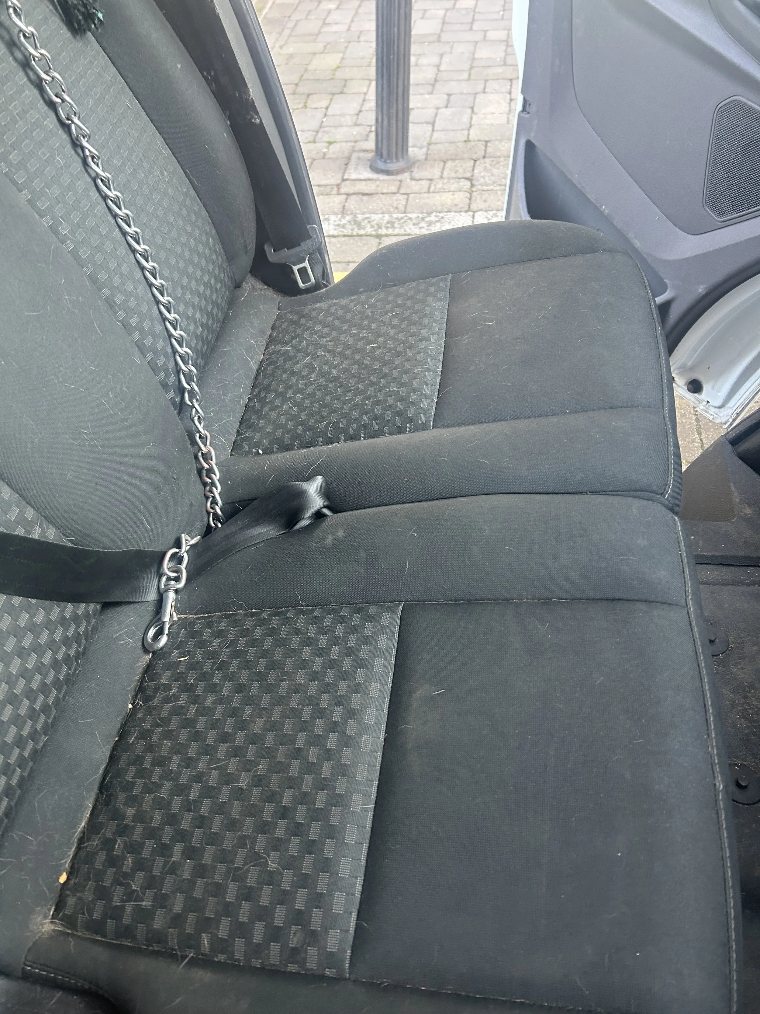 The back seat of a vehicle with black fabric seats, some visible c hairs, and a metal chain draped over the seat.
