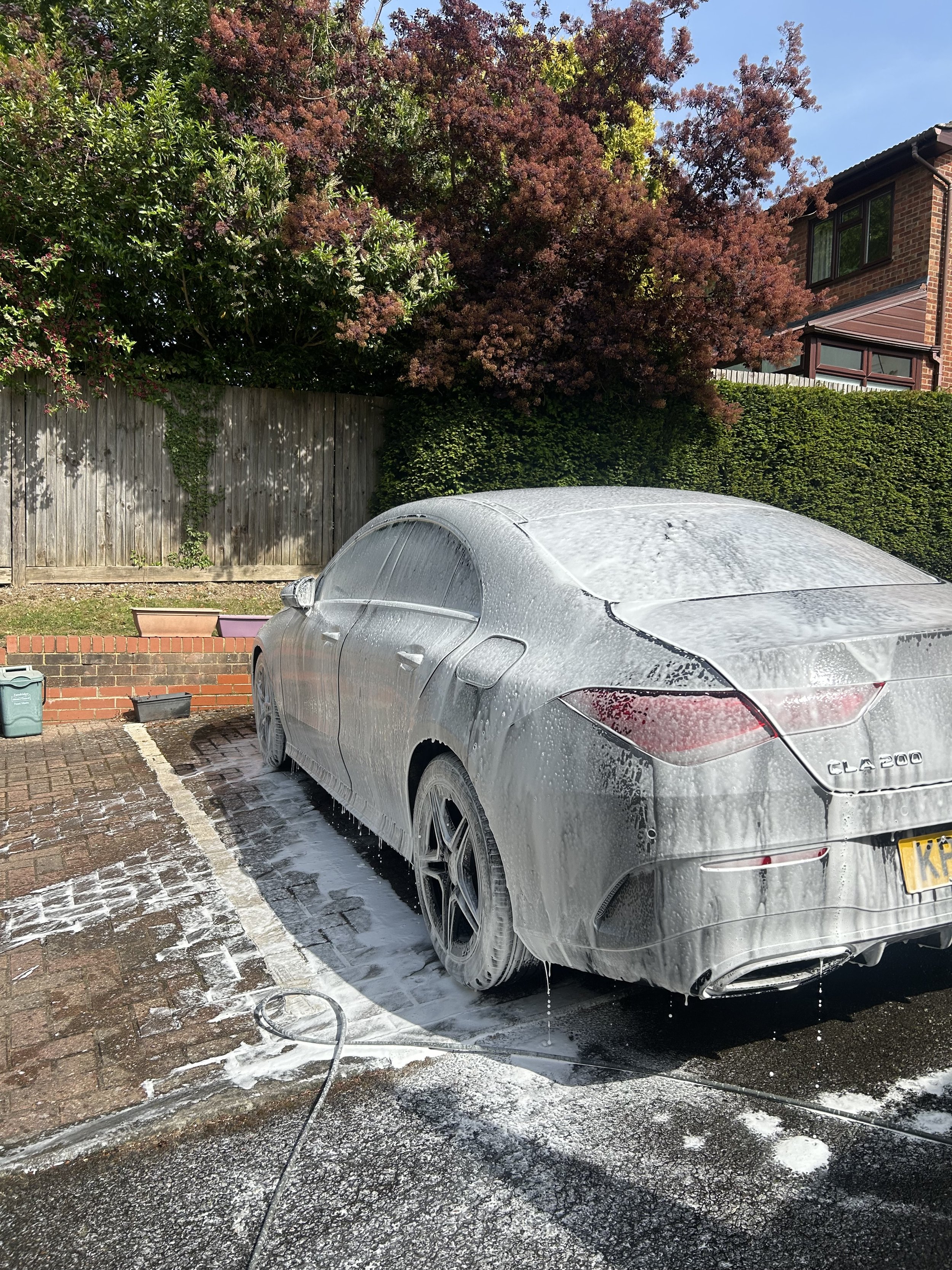 A silver Mercedes-Benz CLA 200 car being washed with soap and water in a parking space. The car is covered in soap suds, and a hose is lying on the ground. The background includes a wooden fence, bushes, and trees with purple and green leaves, and a 