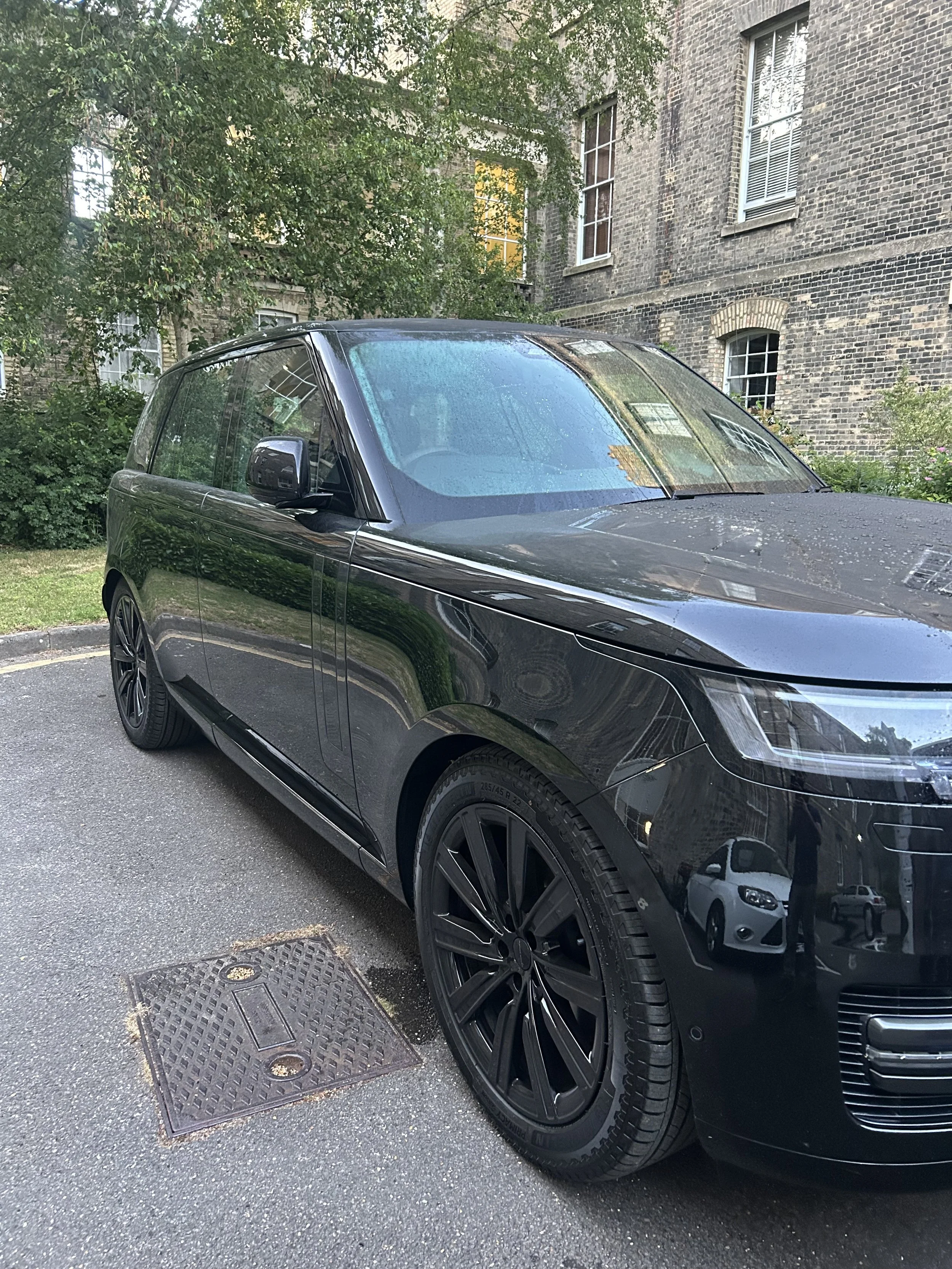 Close-up of a sleek, black luxury car parked on a street near a brick building and some greenery. The car has shiny black wheels and tinted windows.
